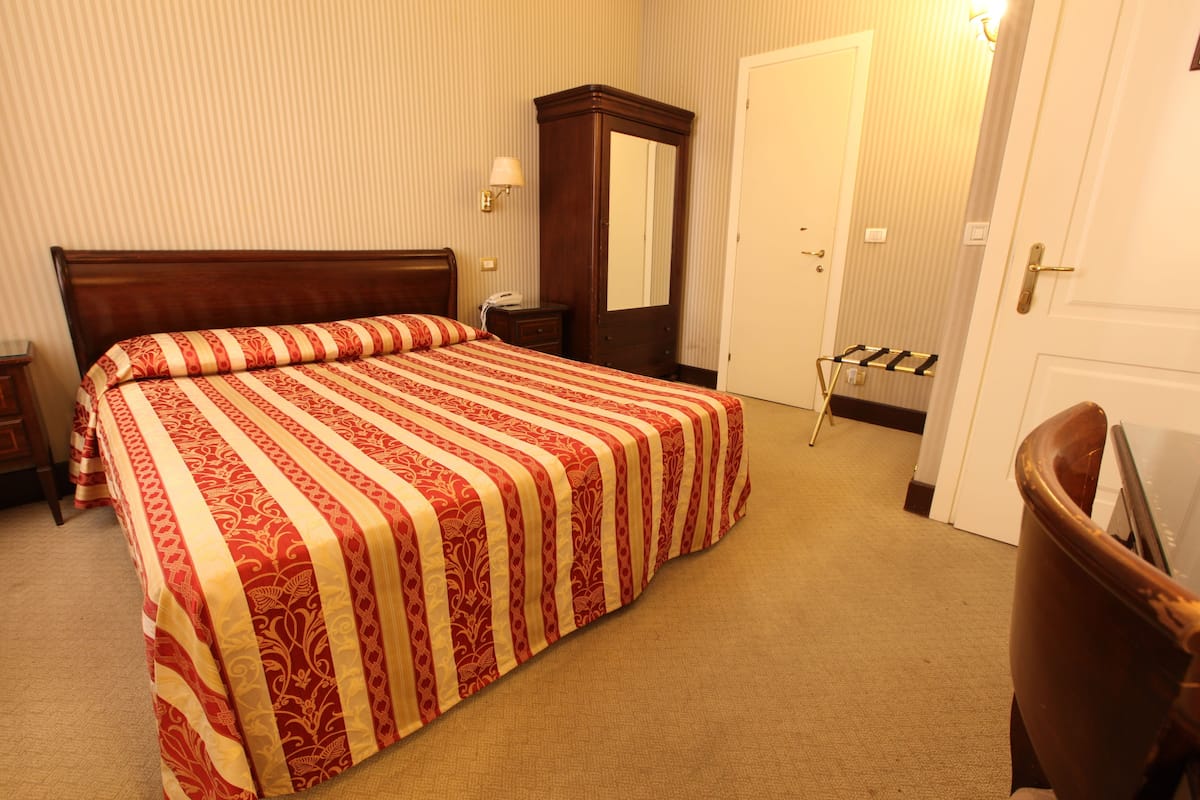 Una cama bien hecha con una colcha a rayas, una mesita de noche de madera con una lámpara, un espejo y una puerta con un perchero para toallas.