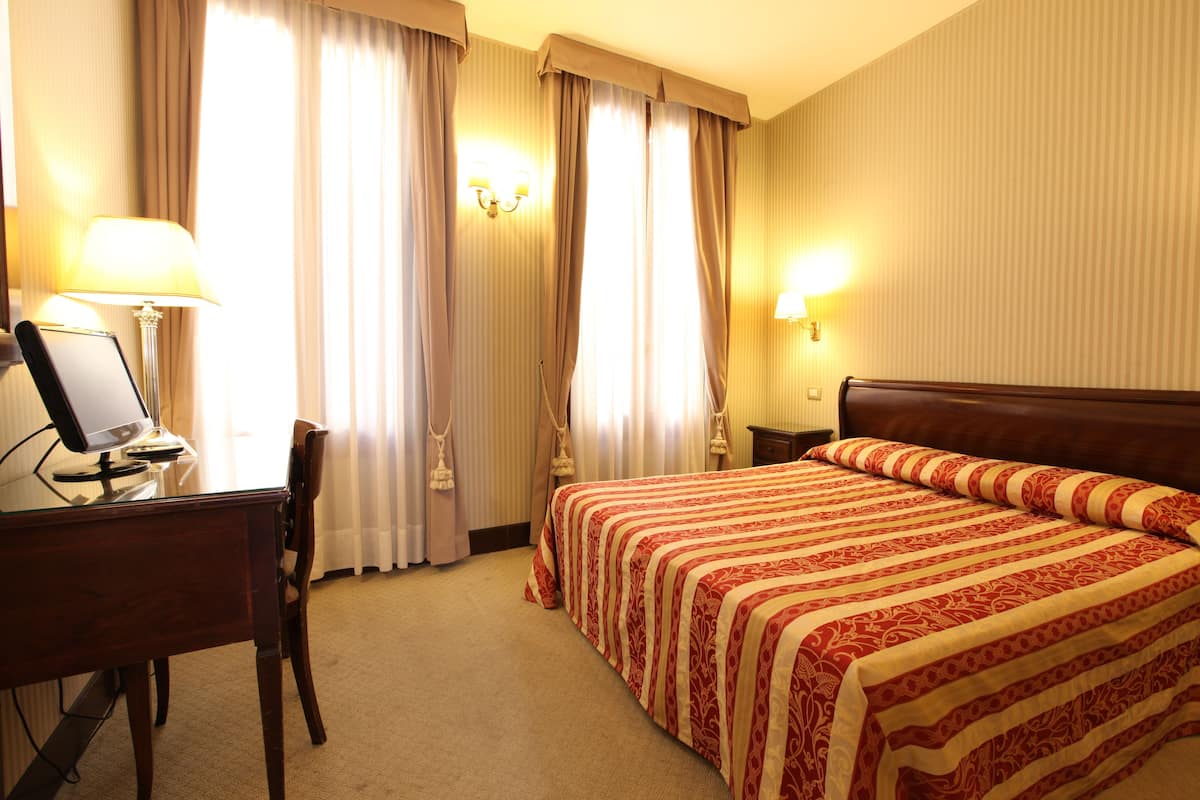 Una habitación de hotel con cama, escritorio con computadora, silla y cortinas.