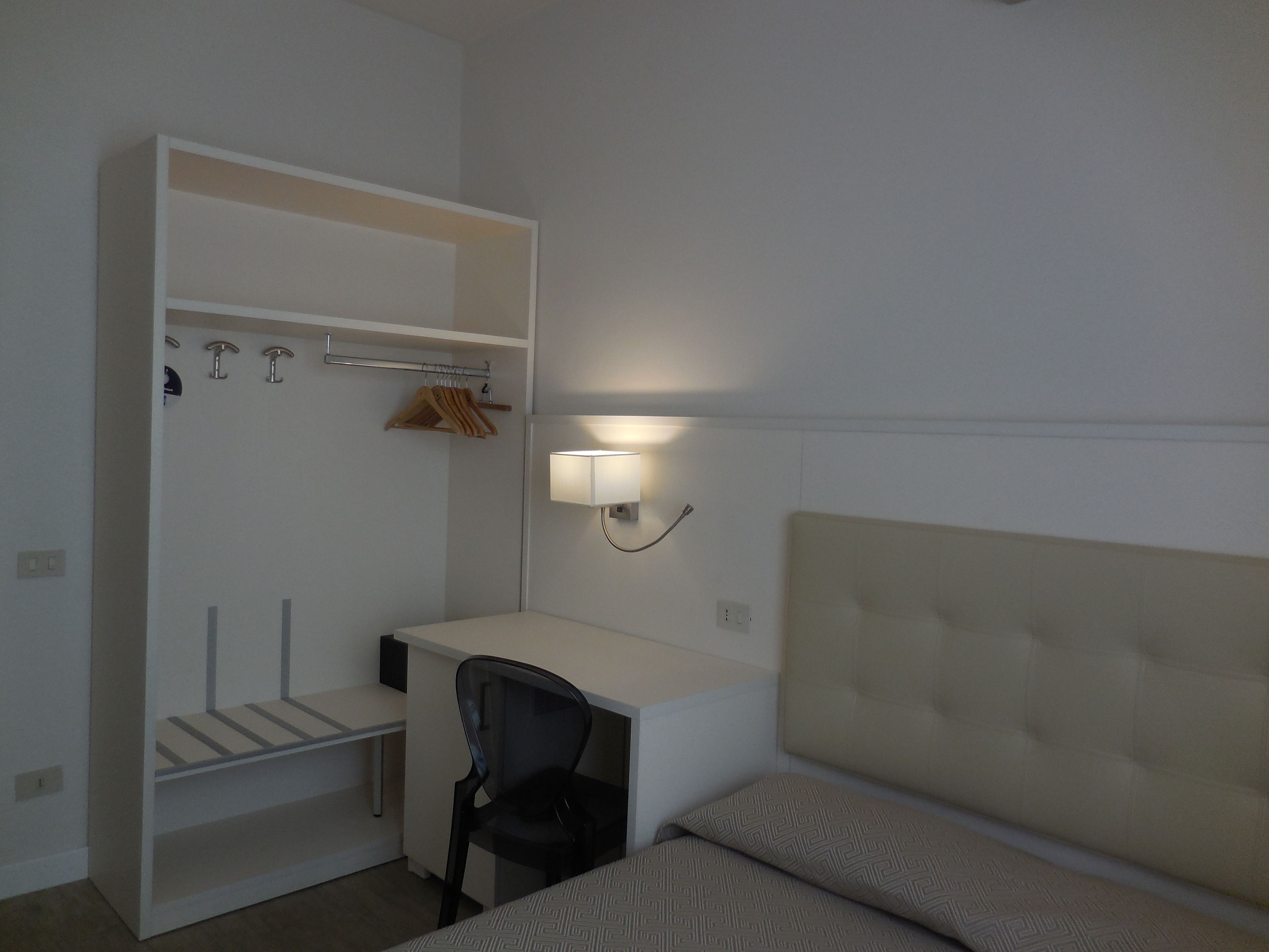 Una camera da letto con un letto, una scrivania e una sedia. Si nota un armadio pieno di vestiti appesi e uno scaffale.
