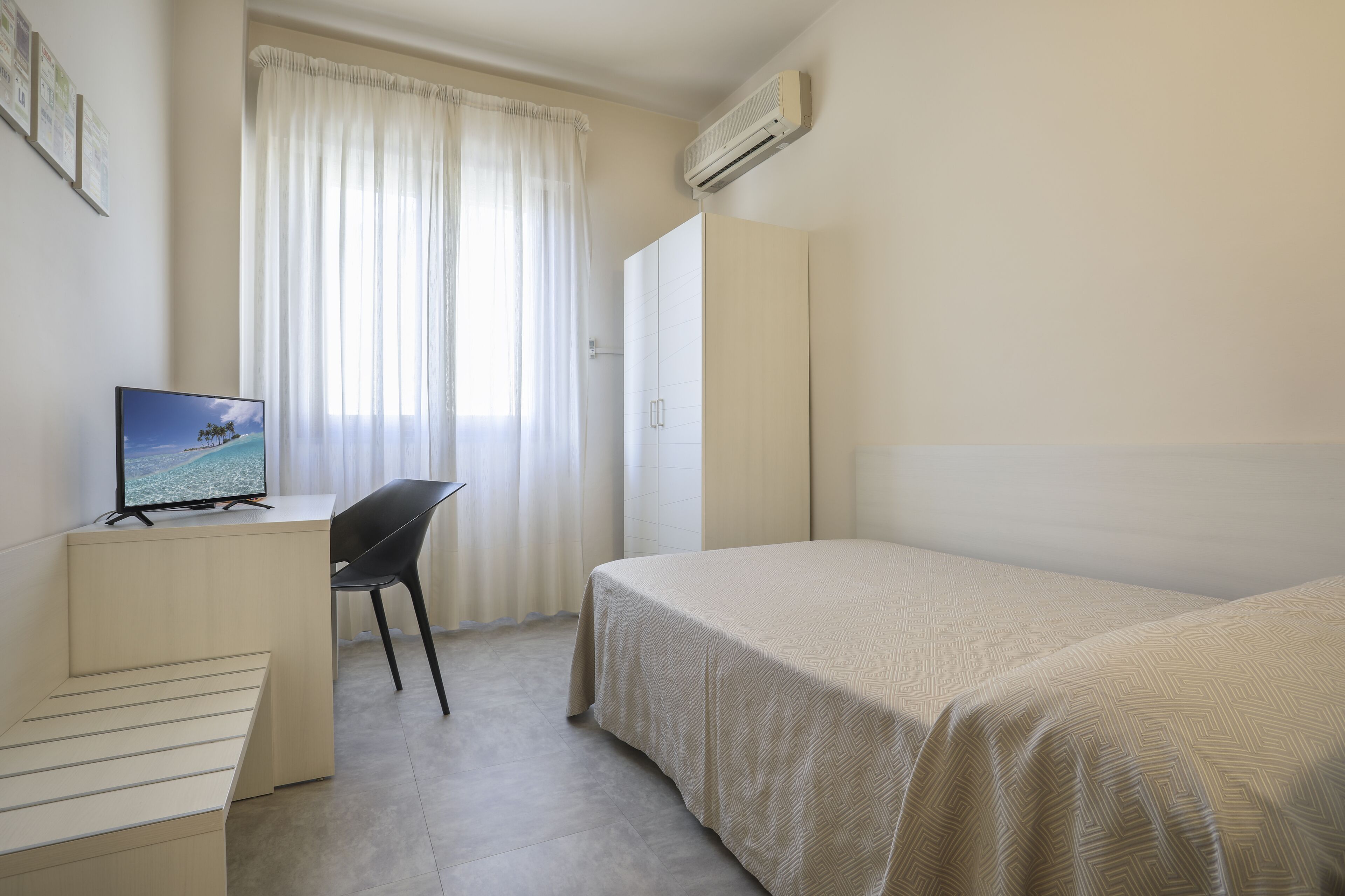 A bedroom with a bed, a desk with a computer, a chair, and a window with curtains.