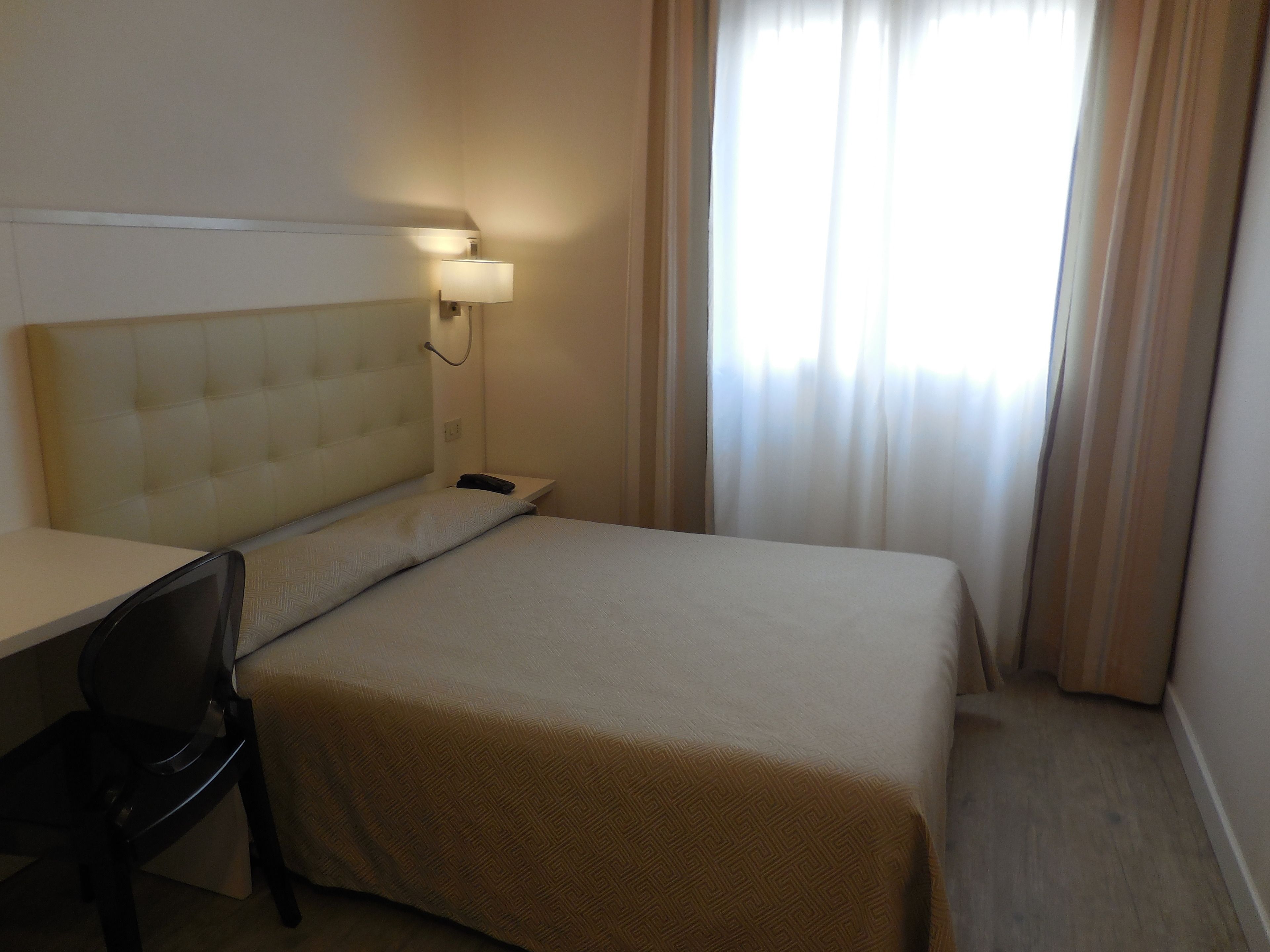 A hotel room with a bed, a desk, a chair, a lamp, and a window with curtains.