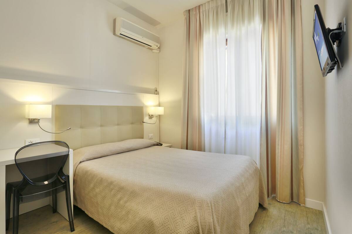Et hotelværelse med en seng, et skrivebord med stol, et fjernsyn ophængt på væggen og en aircondition.