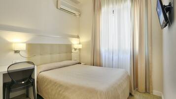 A hotel room with a bed, a desk with a chair, a TV mounted on the wall, and an air conditioning unit.