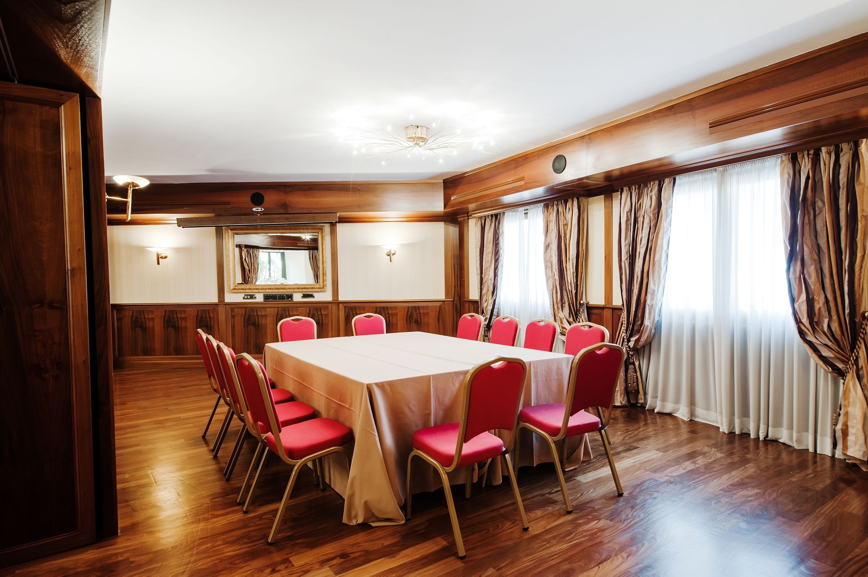 Un salón de conferencias con una mesa grande y sillas rojas, paneles de madera y un ventanal con cortinas.