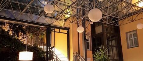 Un atrio con techo de vidrio, una escalera, mobiliario de mimbre y luces esféricas colgantes.