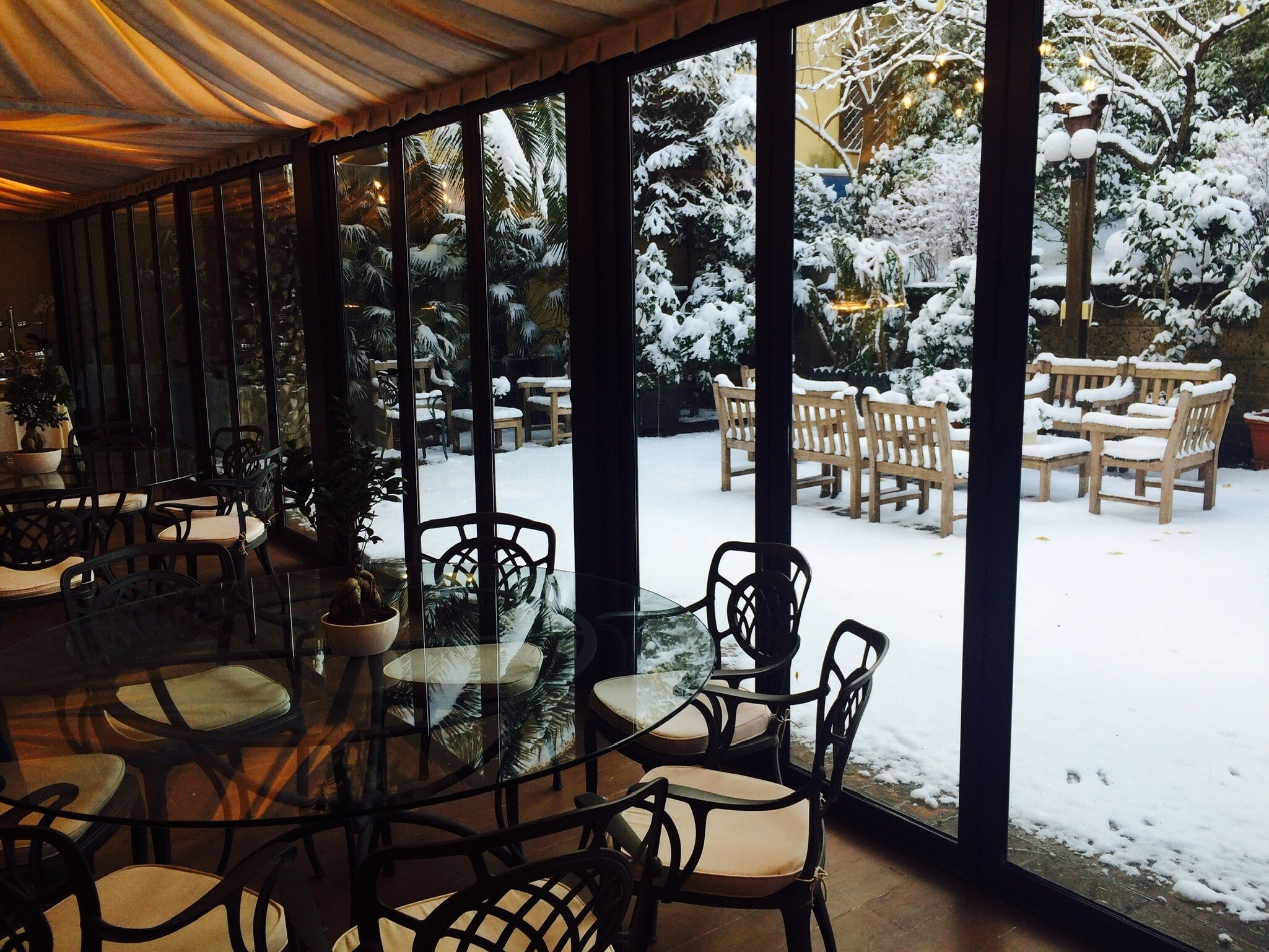 A covered outdoor seating area with glass tables and chairs, overlooking a snowy garden with benches and potted plants.