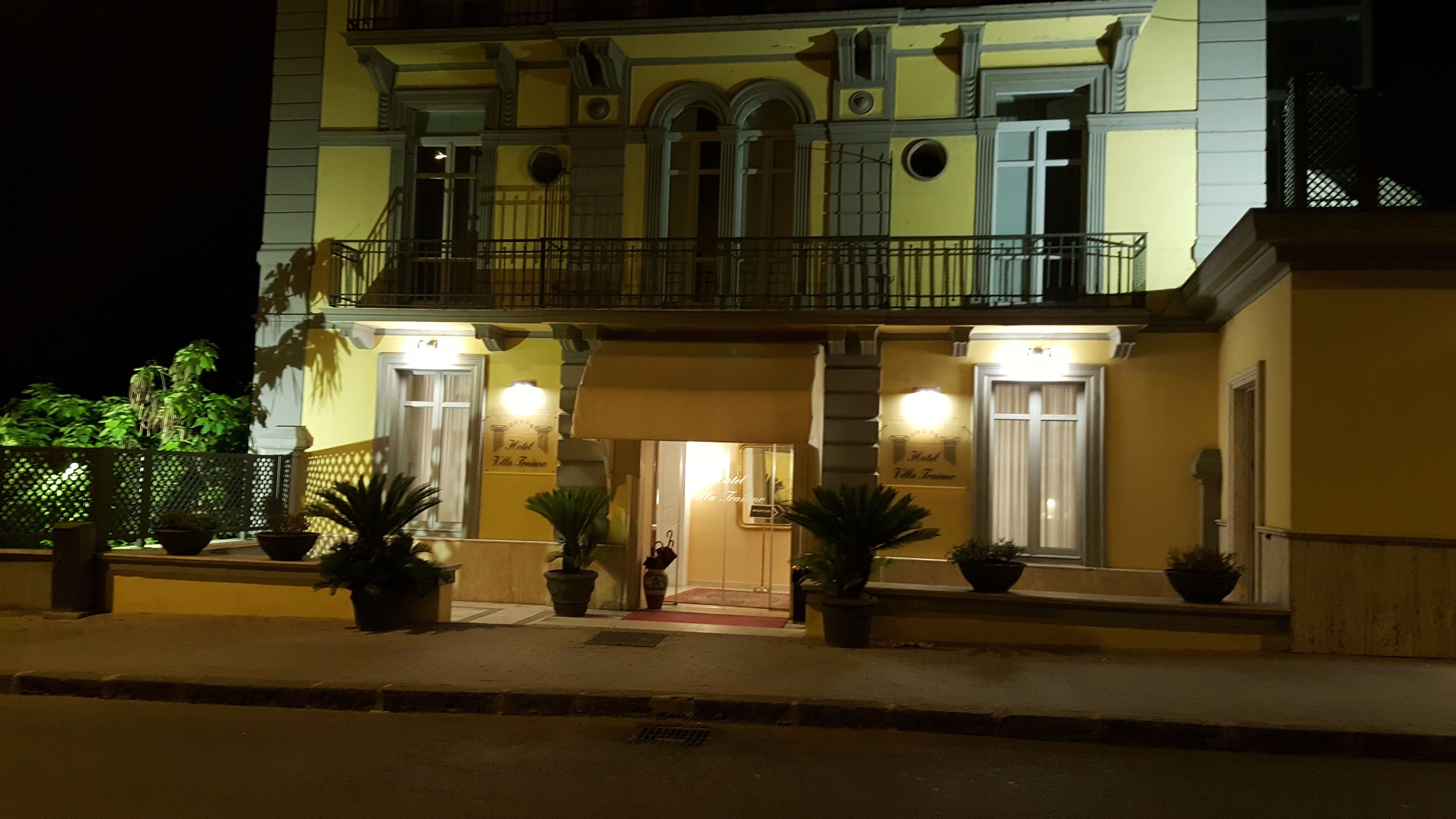 A hotel facade at night with a balcony, potted plants, and illuminated signage.
