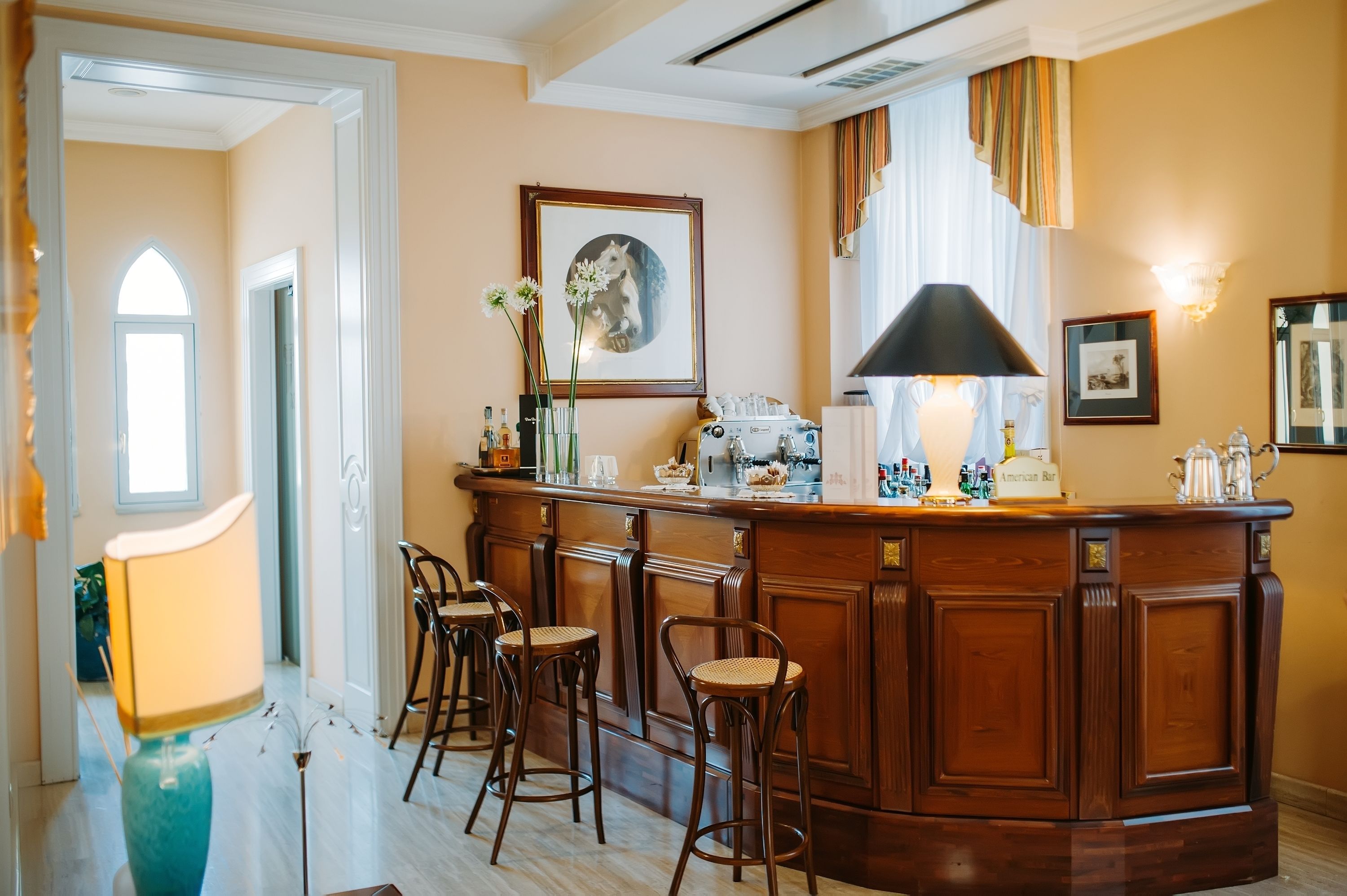 A bar area with a wooden counter, bar stools, a framed picture, and a lamp.