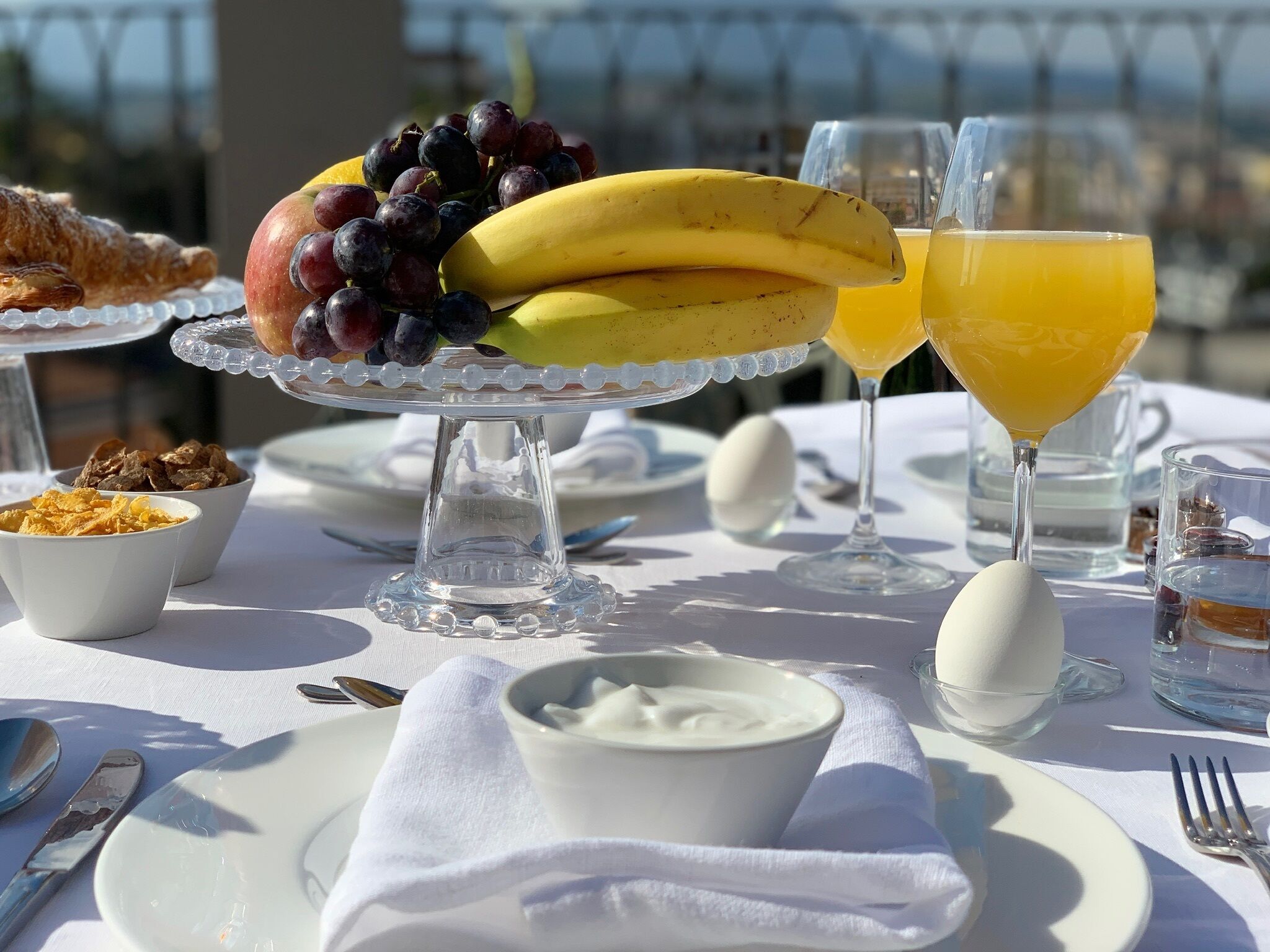 A breakfast setting with a variety of food items including fruits, cereal, and beverages on a table with a view.