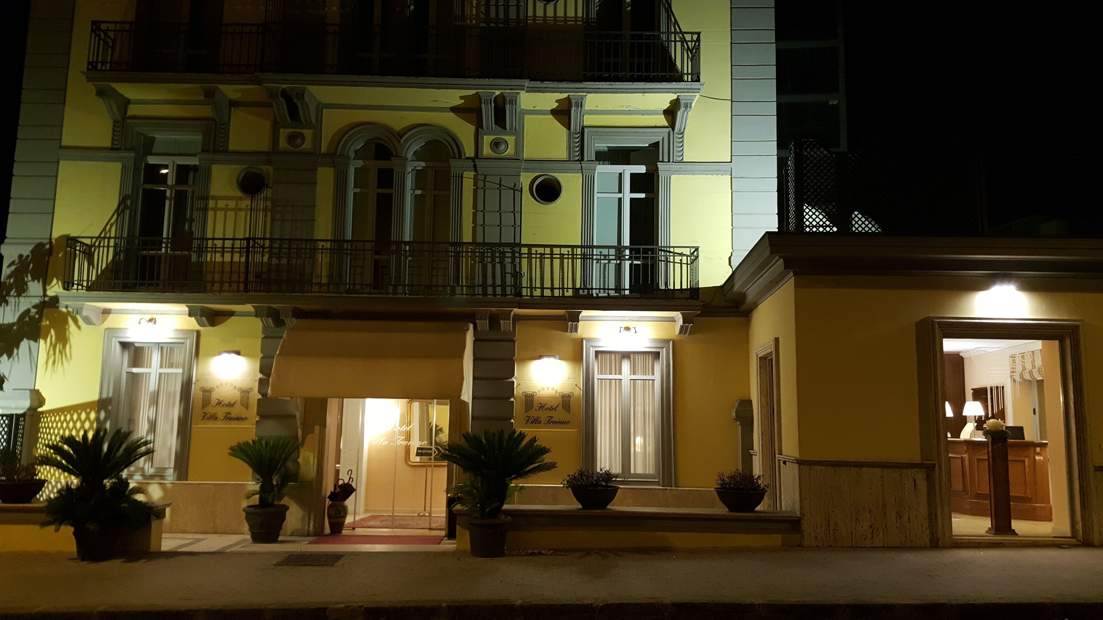 La façade d’un hôtel de nuit, avec des fenêtres et des portes illuminées, un balcon avec des balustrades et des plantes en pot devant.