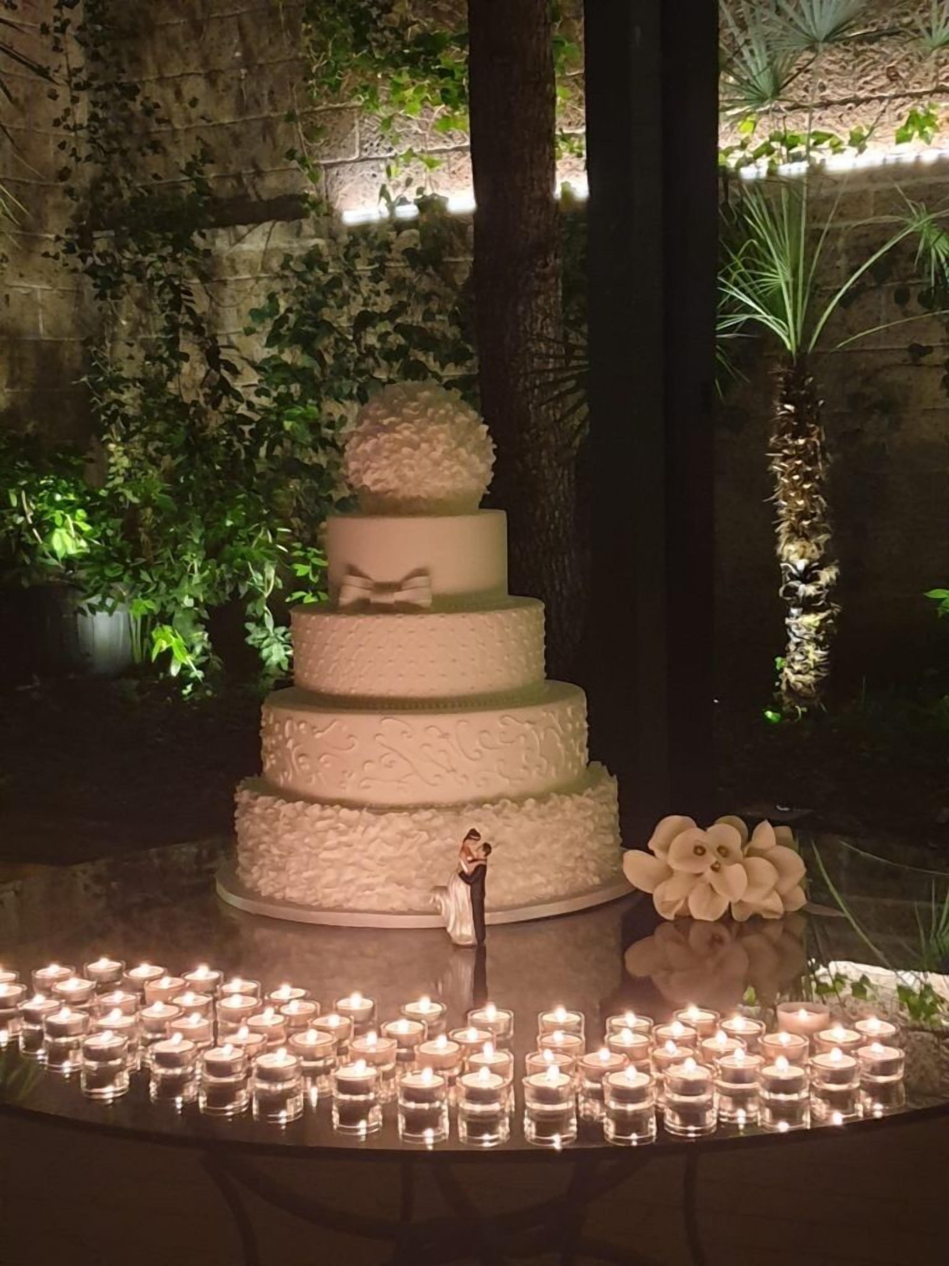 A multi-tiered wedding cake with a figurine topper, surrounded by lit candles, set against a stone wall with greenery.