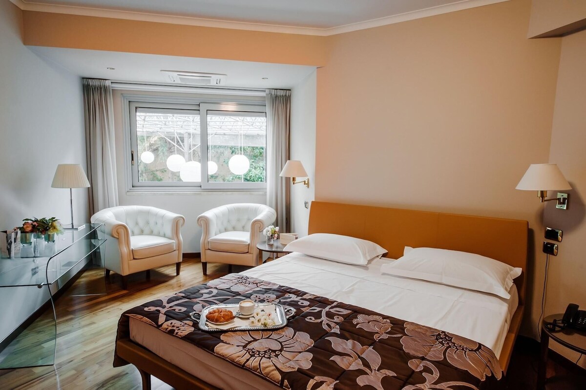 A hotel room with a bed, two chairs, a desk, and a window with curtains.