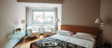 A hotel room with a bed, two chairs, a glass console table, a bedside table, and a window with curtains.