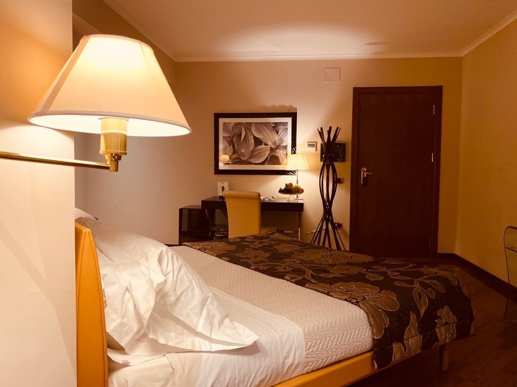 A hotel room with a bed, a desk, a chair, a lamp, a framed artwork, and a door.