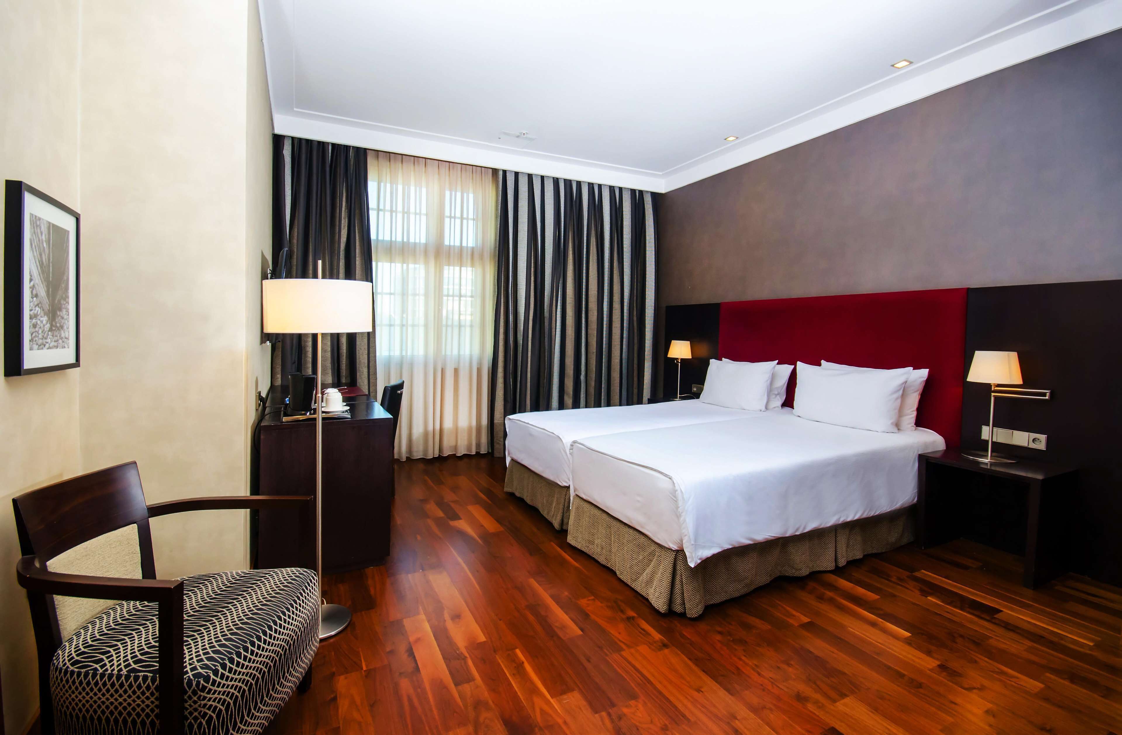 Habitación de hotel con piso de madera, cama con ropa blanca, cabecera roja, dos mesitas de noche con lámparas, un escritorio con una silla y un ventanal con cortinas.