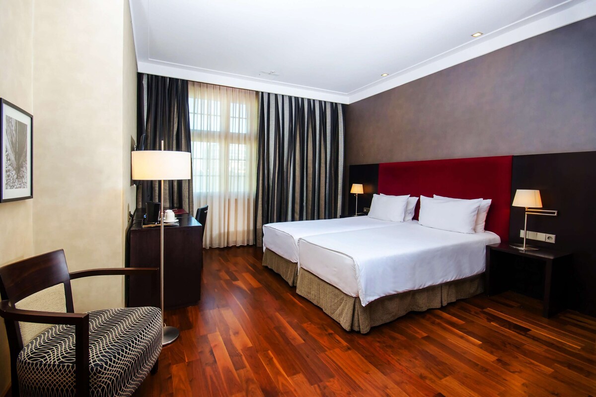 Une chambre d’hôtel avec un plancher en bois, un lit recouvert de draps blancs, une tête de lit rouge, deux tables de chevet avec des lampes, un bureau avec une chaise et une grande fenêtre avec des rideaux.