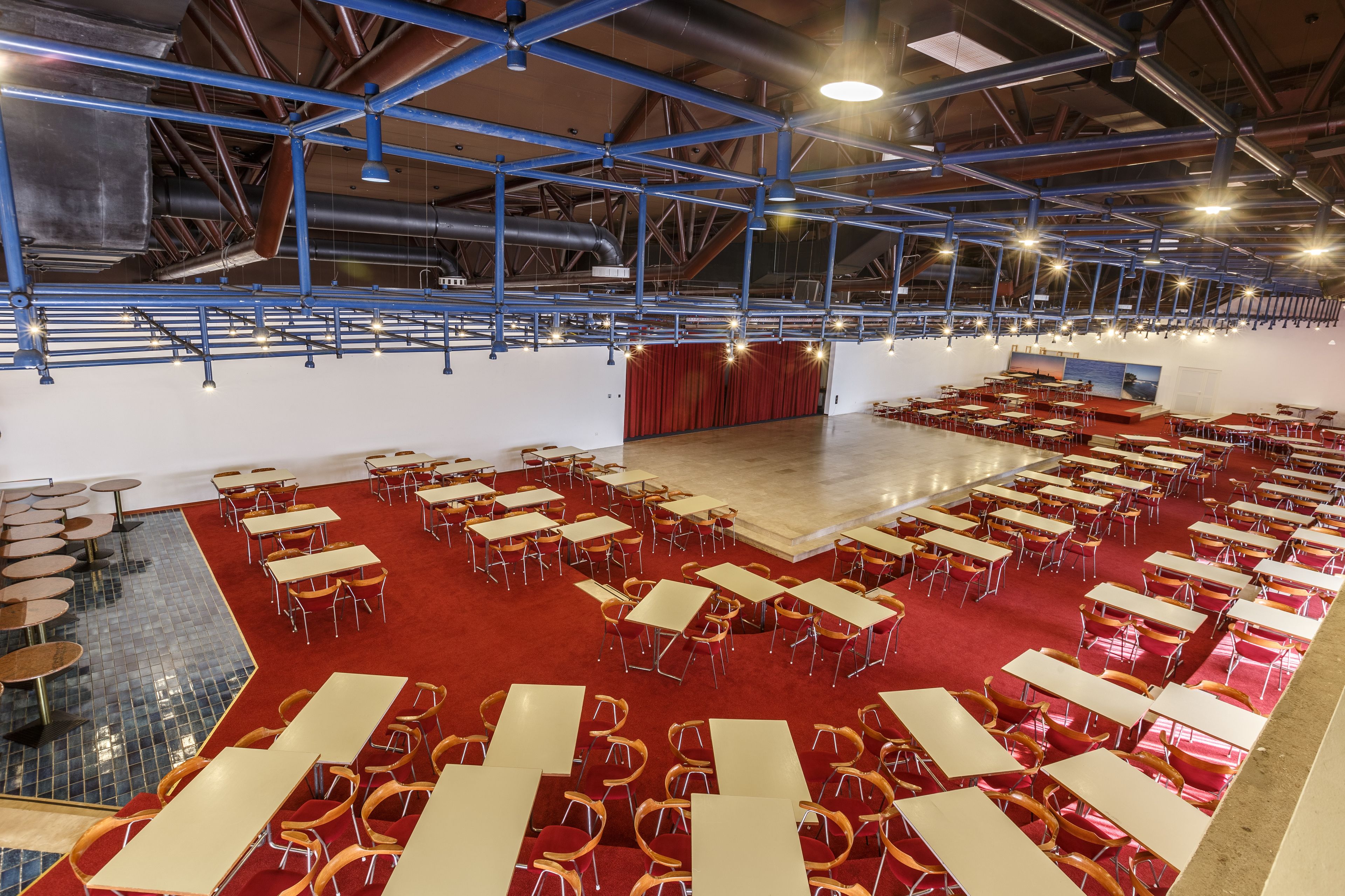 Een ruime binnenruimte met meerdere tafels en stoelen in rijen, een podium met rode gordijnen, en een hoge plafond met zichtbare balken en verlichtingsarmaturen.
