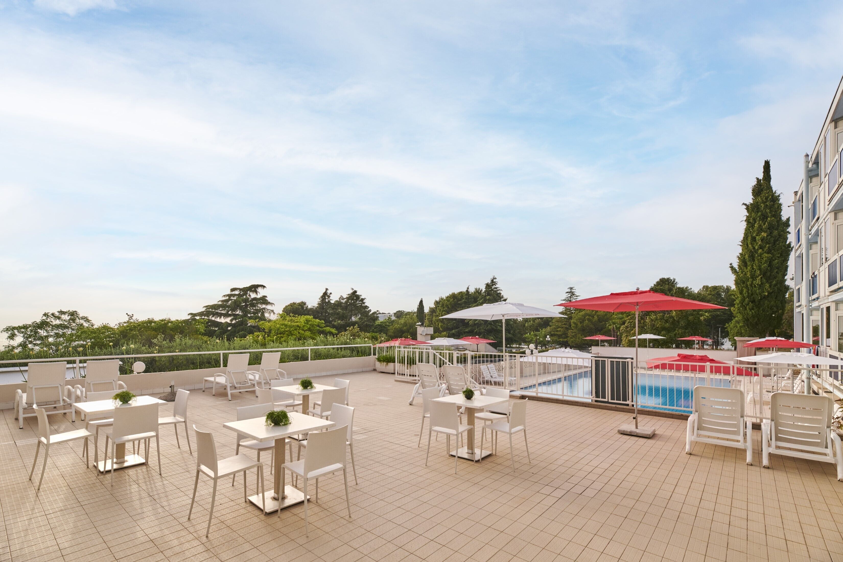 A rooftop terrace with white tables and chairs, red umbrellas, and a pool.