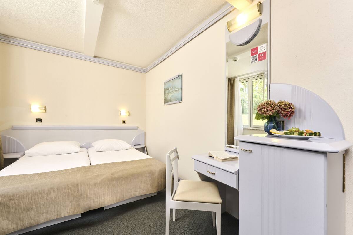 Hotellihuone, jossa on sänky, työpöytä, tuoli, peili ja maljakko, jossa on kukkia.