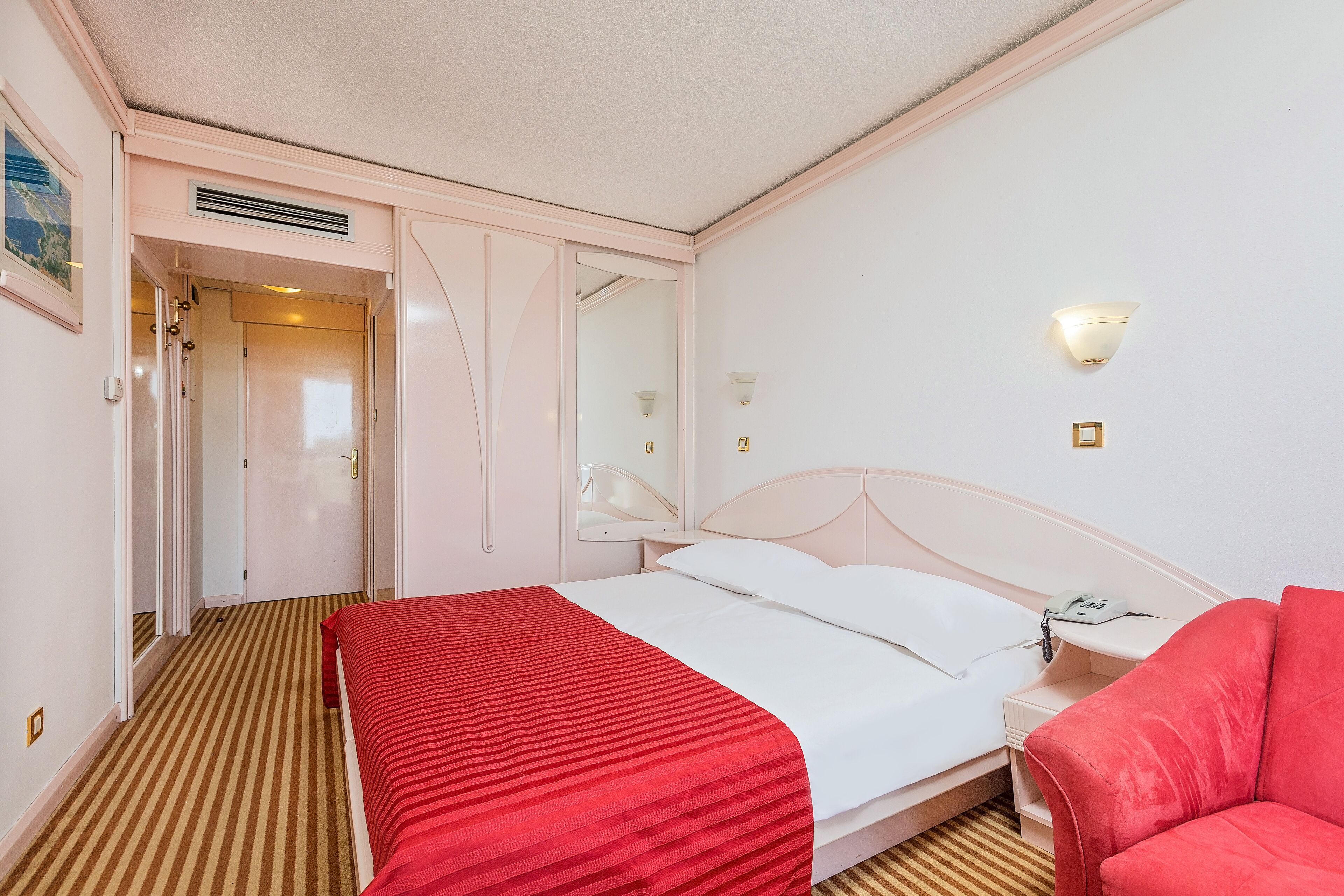 Una camera d'albergo con un letto, un tappeto a righe rosse e bianche, un comodino bianco con un telefono, una sedia rossa e un lampadario a parete.