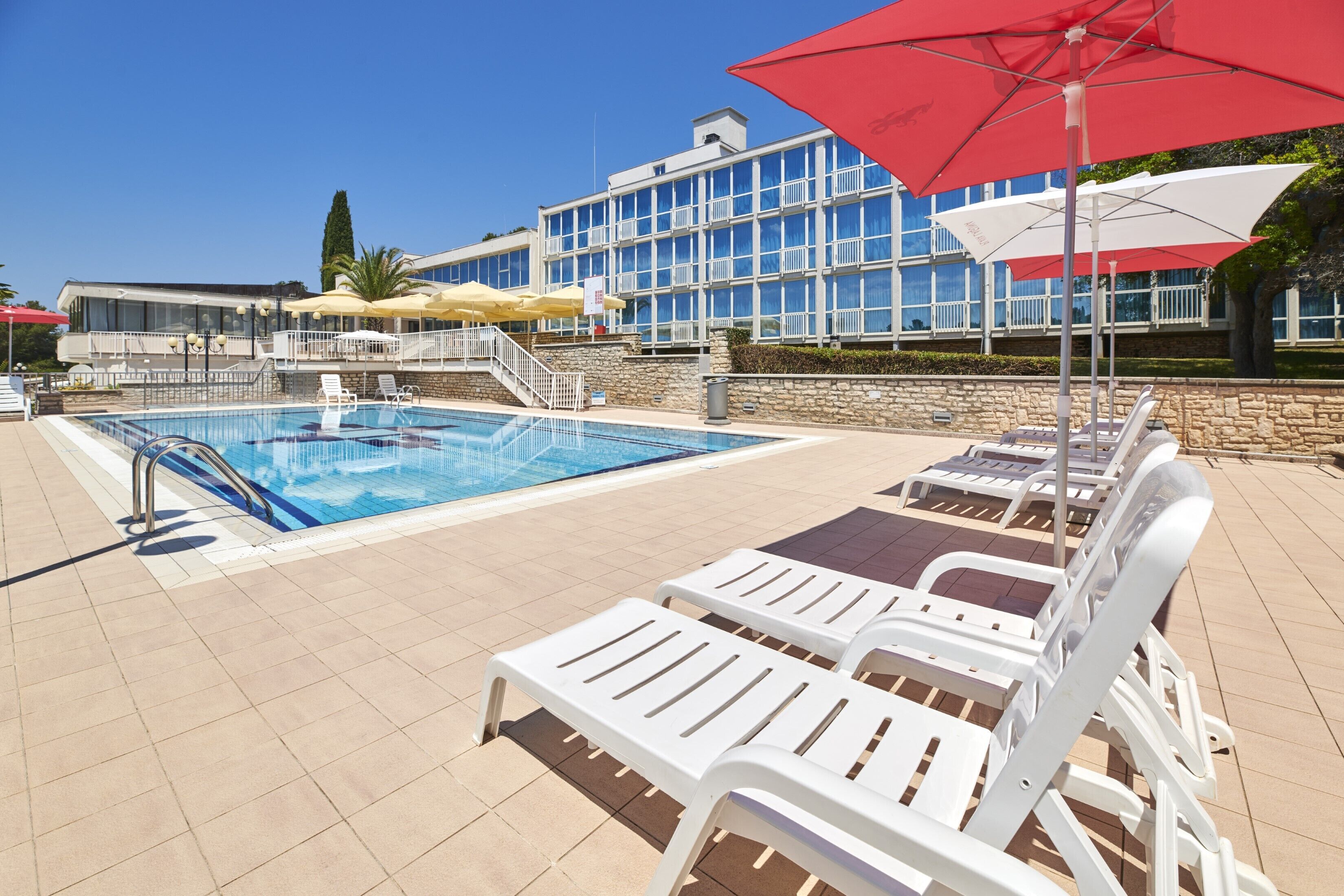 A swimming pool with lounge chairs and umbrellas, a stone wall, and a building with balconies in the background.