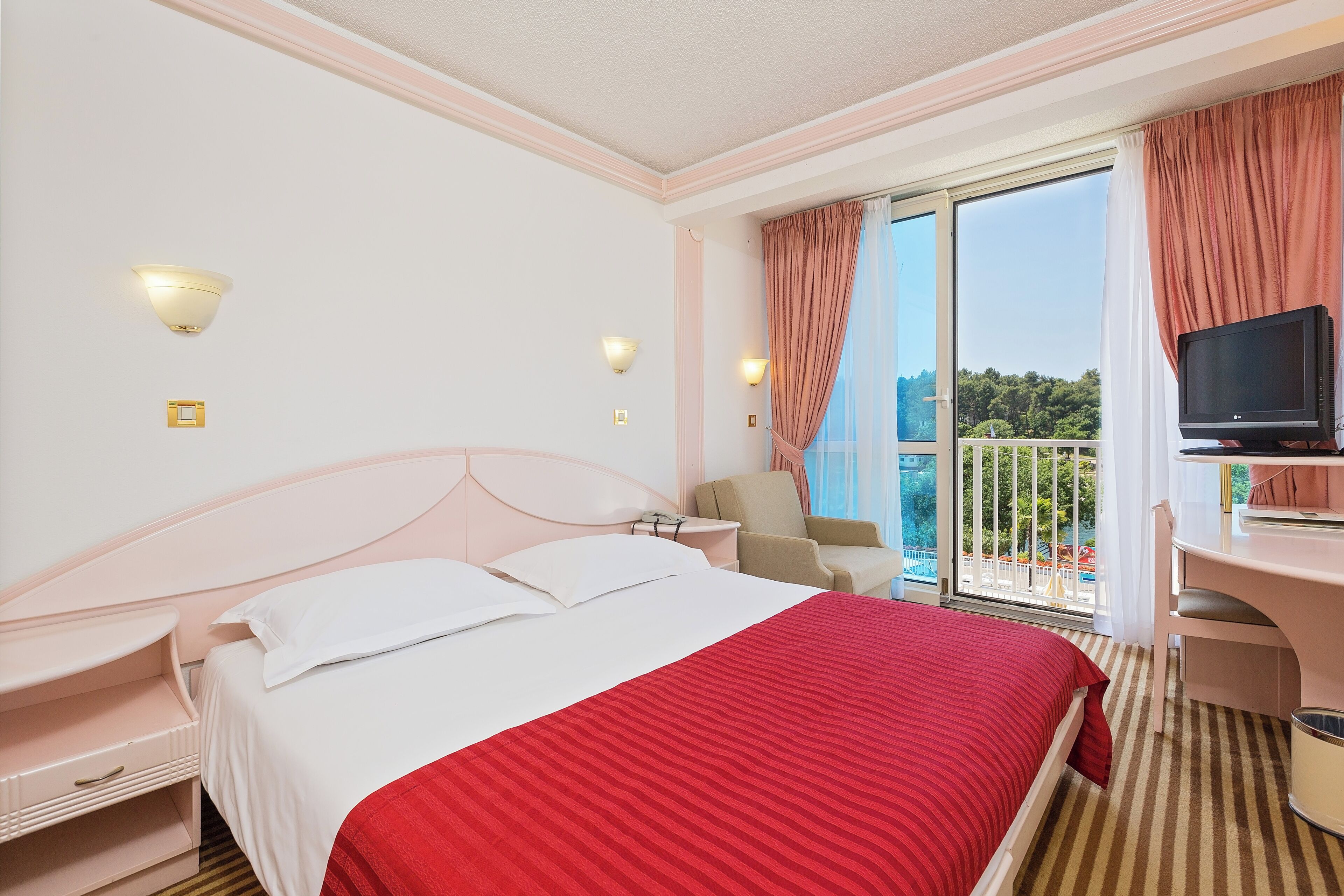 Une chambre d’hôtel équipée d’un lit, d’un bureau, d’une chaise, d’une télévision et d’un balcon avec vue.