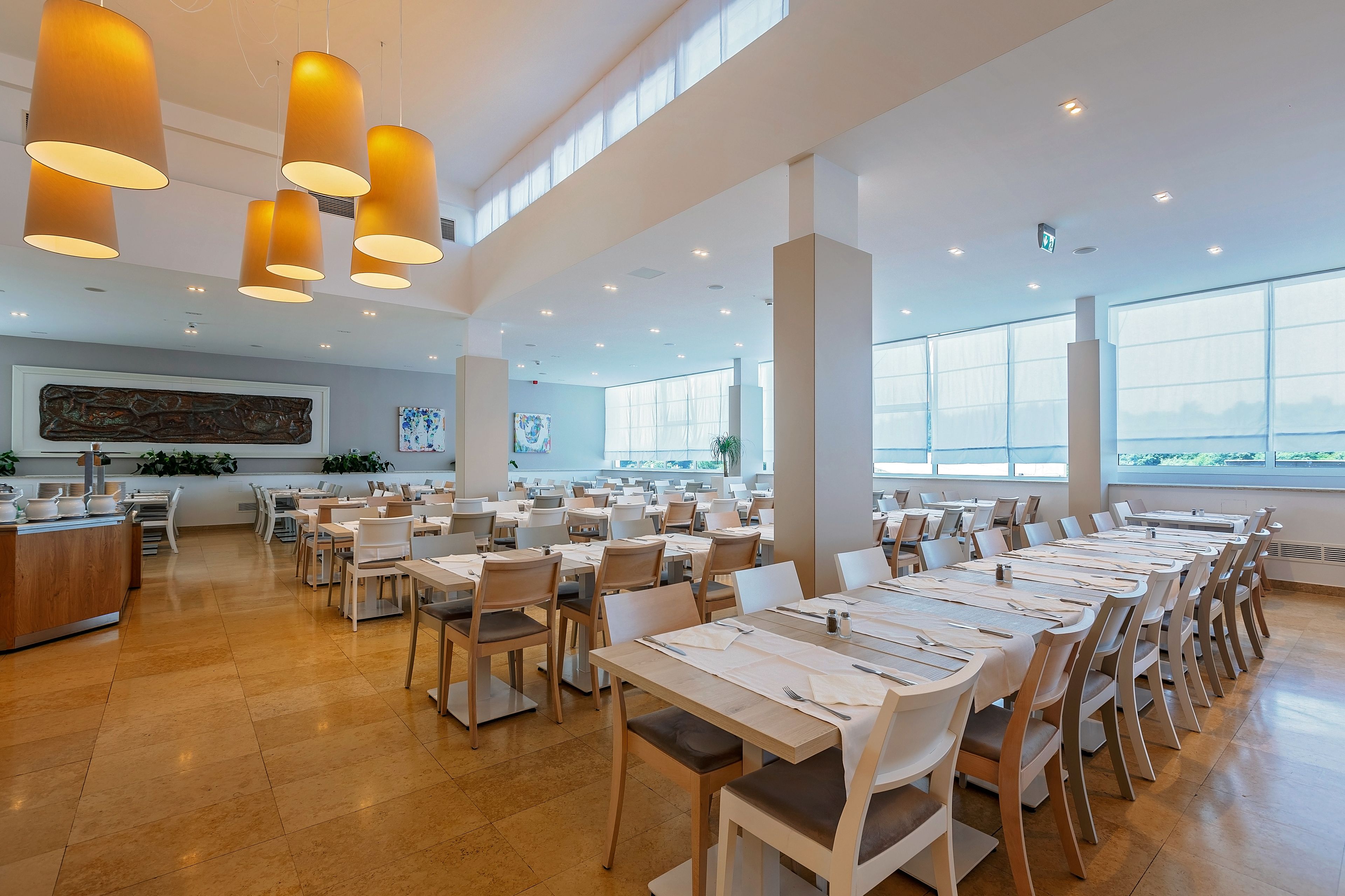 A spacious dining area with multiple tables and chairs, large windows, and pendant lighting.