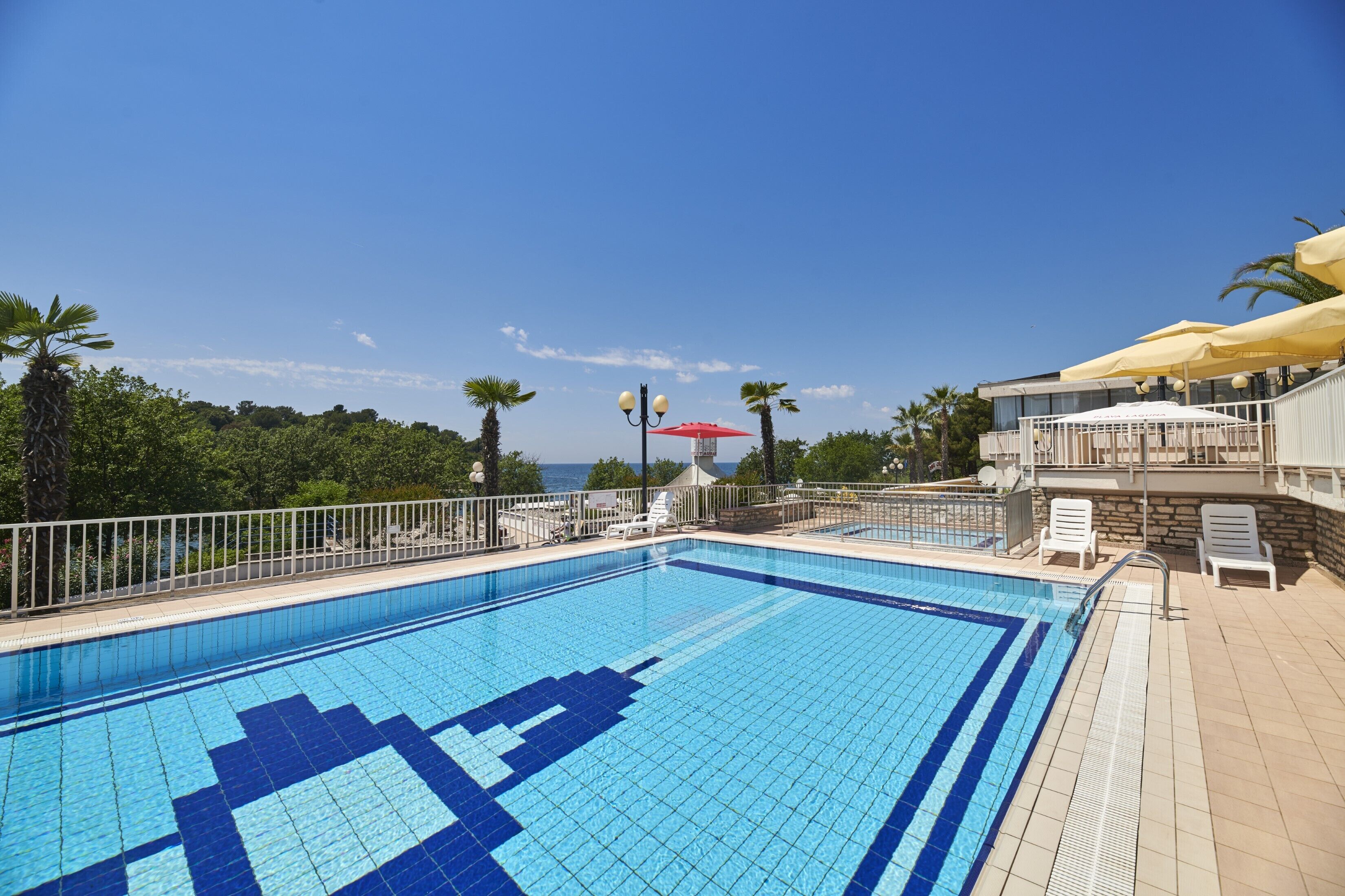 Una piscina con un diseño de mosaicos, rodeada por una cerca blanca y con vistas a un paisaje con palmeras y cielo despejado.