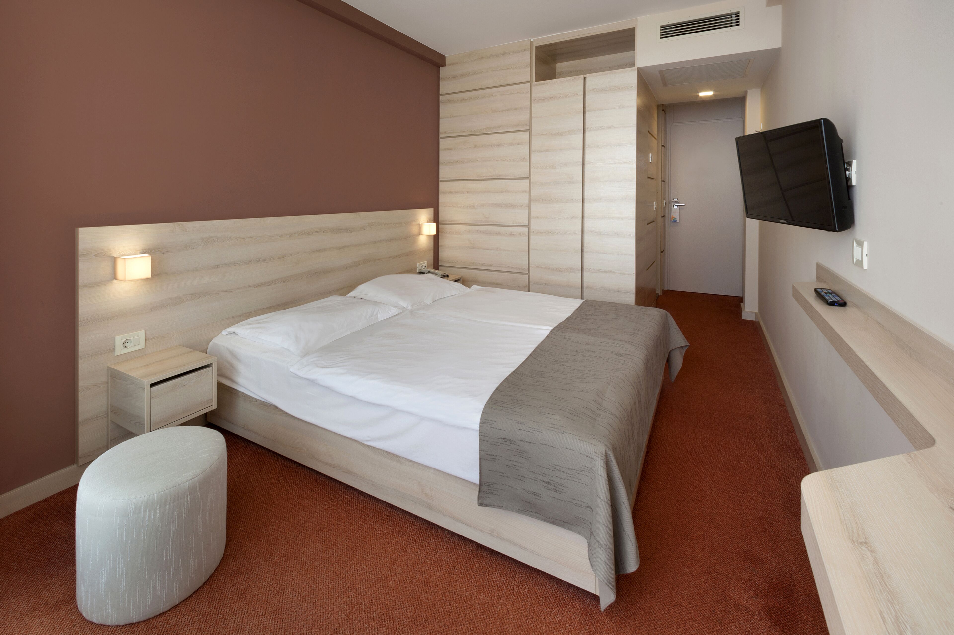A modern hotel room with a large bed, a bedside table, a TV mounted on the wall, and a wardrobe.