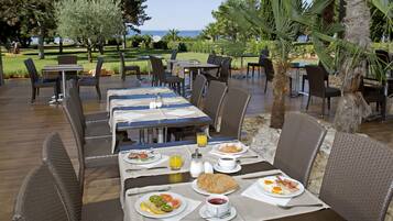 Un área de comedor al aire libre con mesas y sillas, vistas al mar y un jardín bien cuidado.