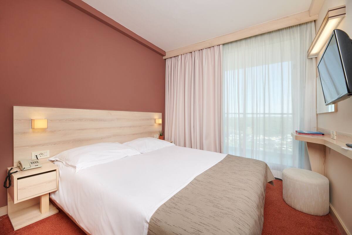 A hotel room with a bed, bedside table, lamp, TV, and a window with curtains.