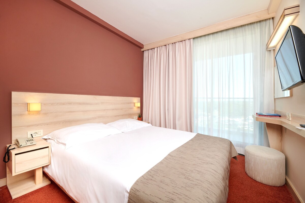 Habitación de hotel con cama, mesita de noche, lámpara, televisor y ventana con cortinas.