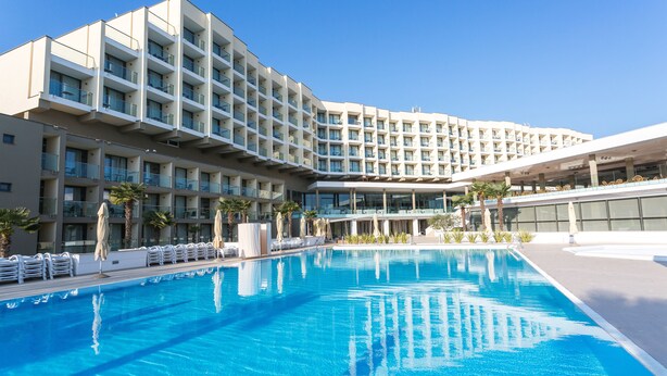 Un hotel moderno con una piscina grande, varios balcones y un cielo azul despejado.
