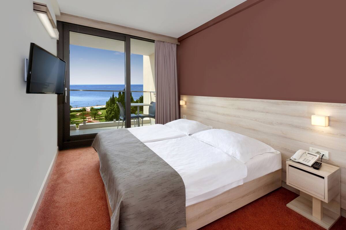 A hotel room with a bed, a nightstand with a phone, a TV mounted on the wall, and a large window offering a view of the sea and greenery.