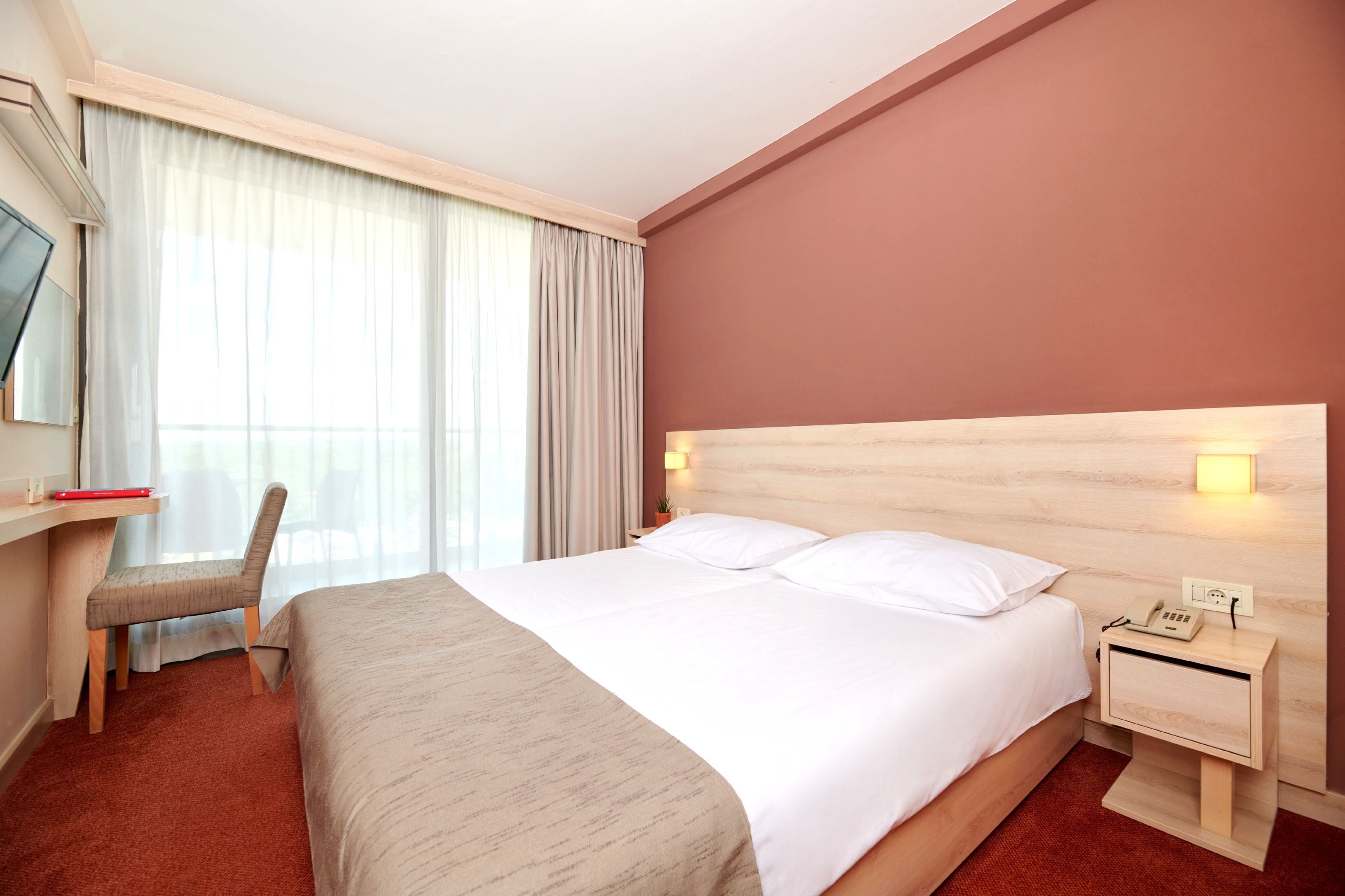 Ett hotellrum med en säng, ett skrivbord, en stol, ett nattduksbord med en telefon och ett fönster med gardiner.