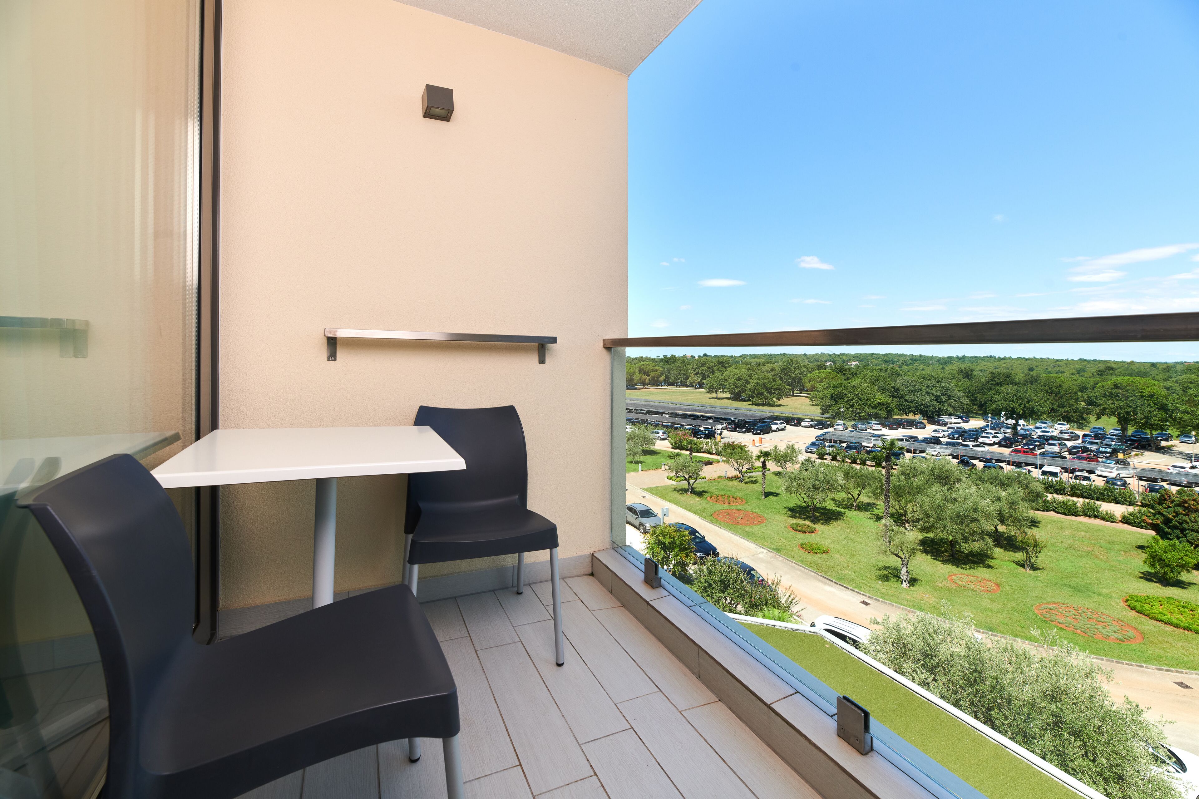 Ein Balkon mit Tisch und Stühlen, einer Glastrennwand und Blick auf einen Parkplatz sowie Grünflächen.