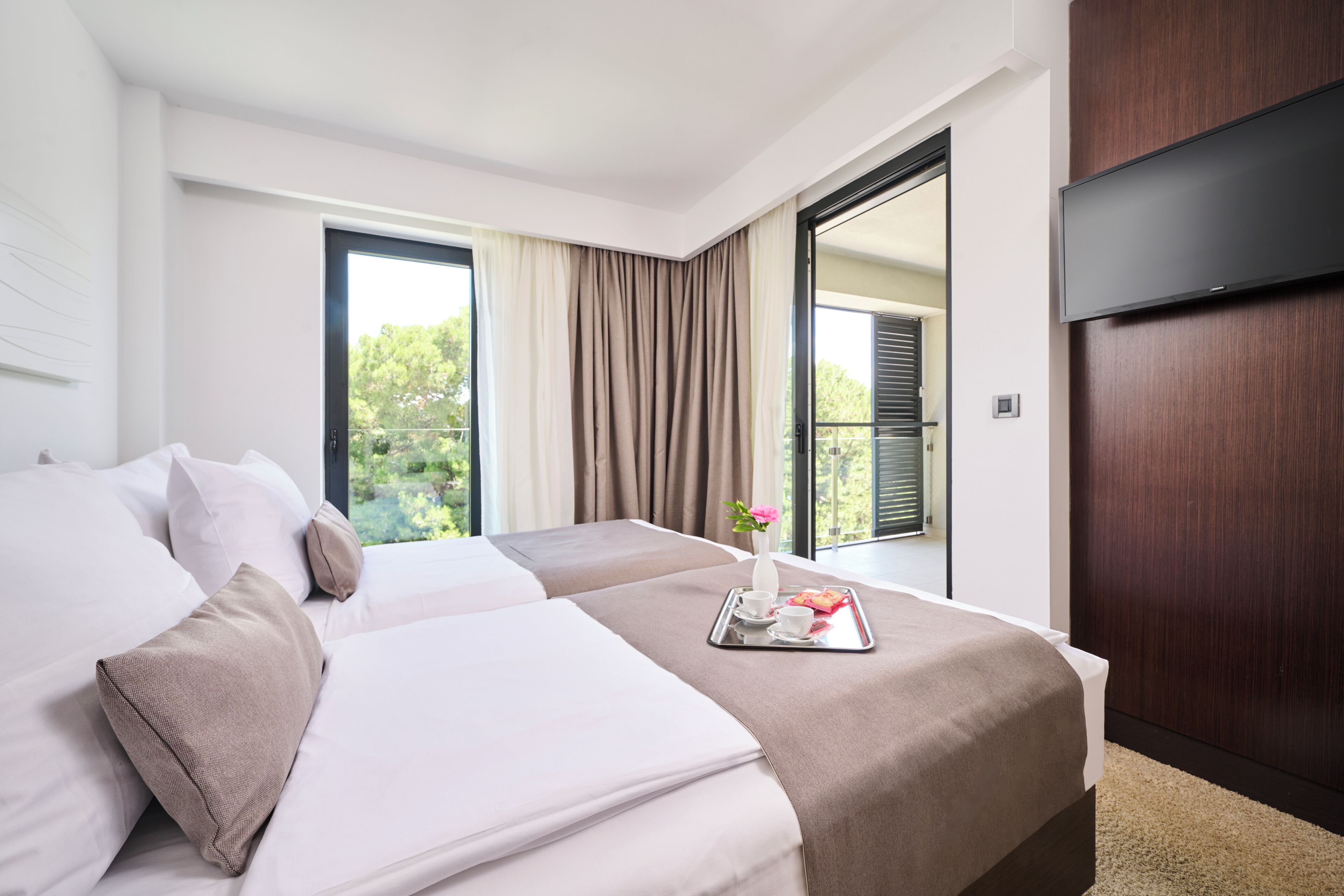 Habitación de hotel con dos camas, un ventanal grande, un televisor de pantalla plana y una bandeja con una flor y un juego de té.