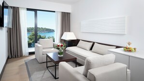 A modern hotel room with a sofa, armchairs, a coffee table, a TV, a lamp, and a view of the sea. - Hotel Parentium Plava Laguna (Porec)