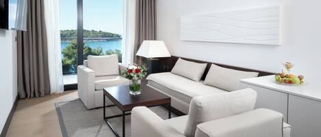 A modern hotel room with a sofa, armchairs, a coffee table, a TV, a lamp, and a view of the sea.