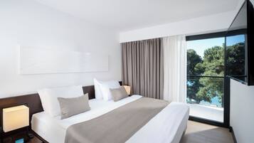 Una habitación de hotel moderna con una cama grande, un televisor de pantalla plana, una mesita de noche con lámpara y vistas a árboles por la ventana.
