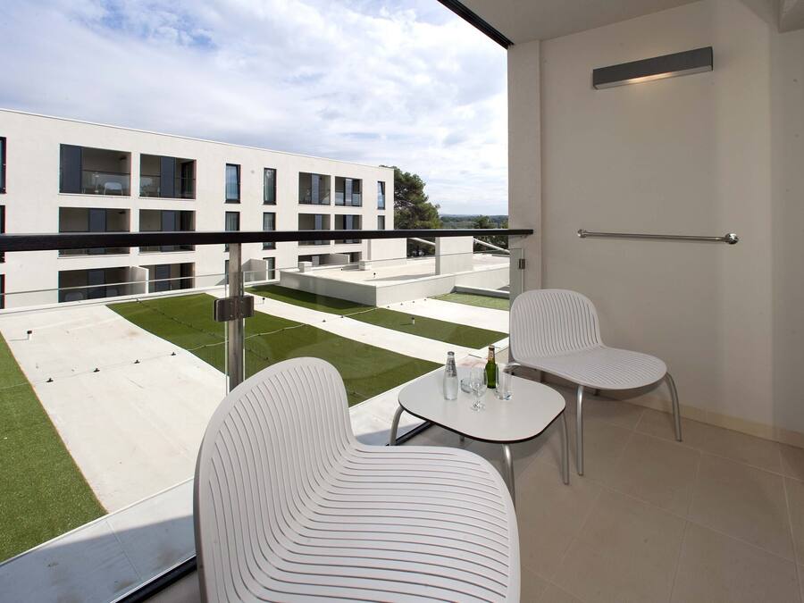 A balcony with two white chairs and a small table, overlooking a building with multiple windows and a grassy area.