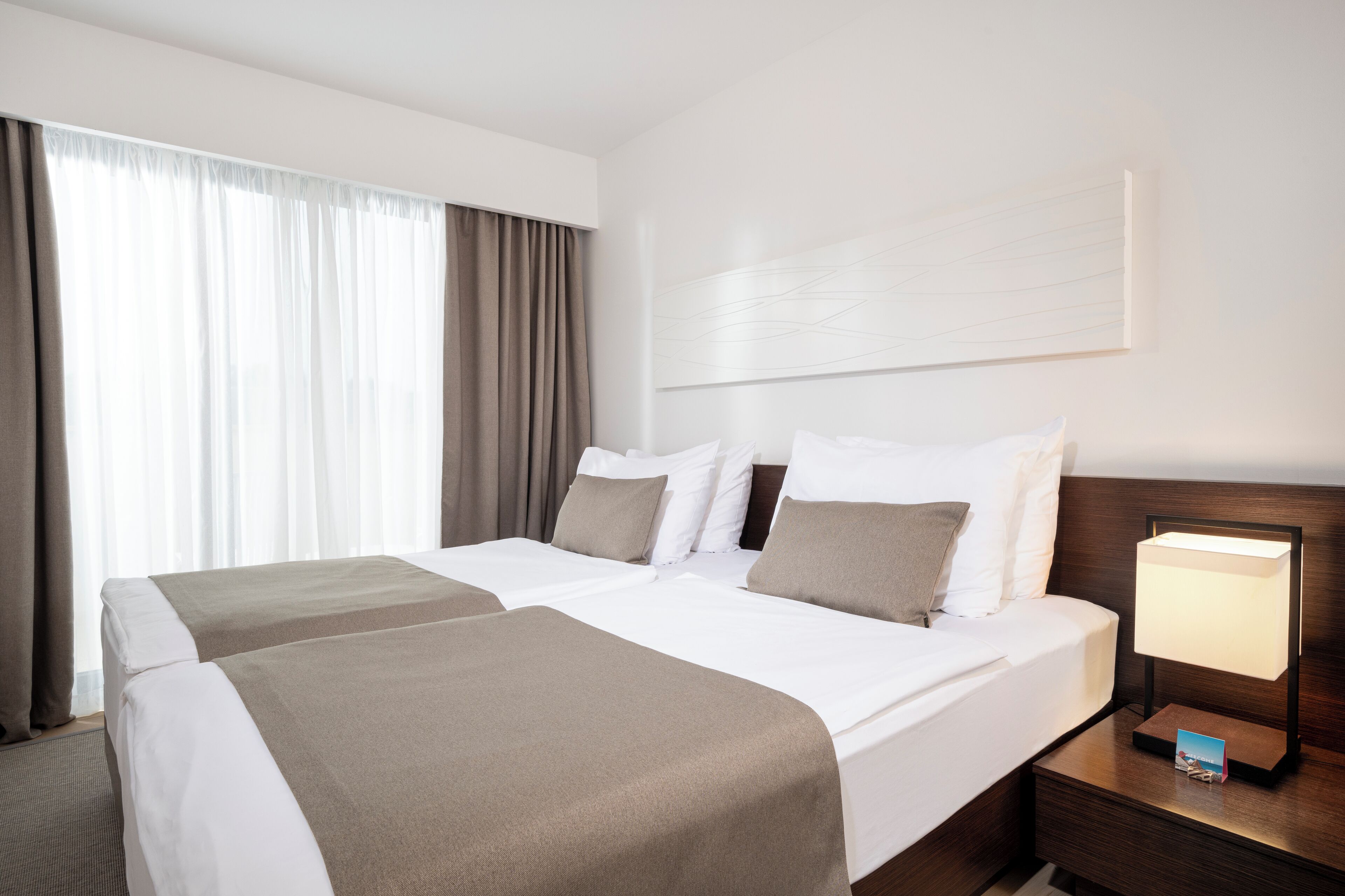 A hotel room with two beds, a nightstand with a lamp, and a large window with curtains.
