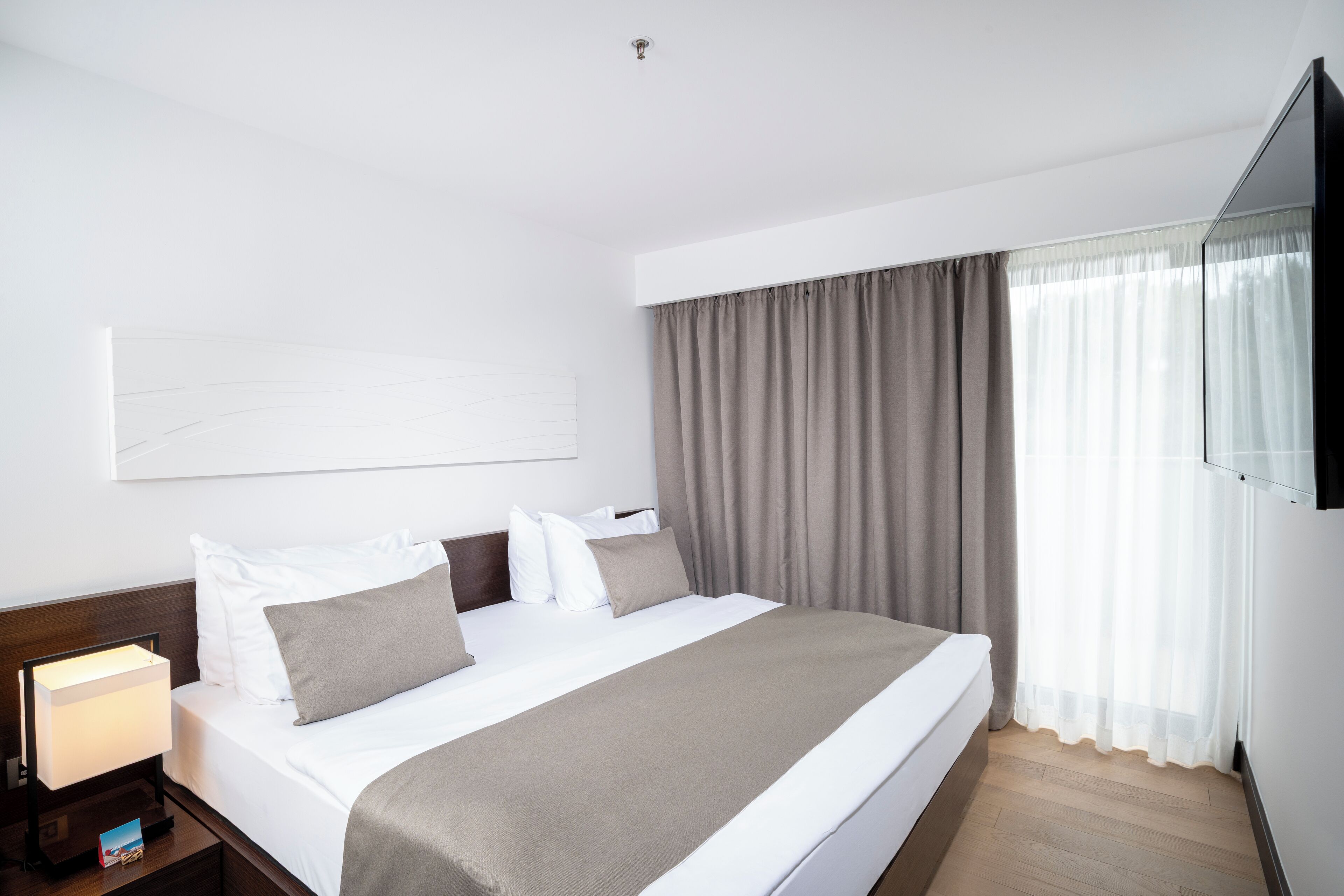 Ein Hotelzimmer mit einem großen Bett, einem Nachttisch mit Lampe, einem Flachbildfernseher und einem großen Fenster mit Vorhängen.