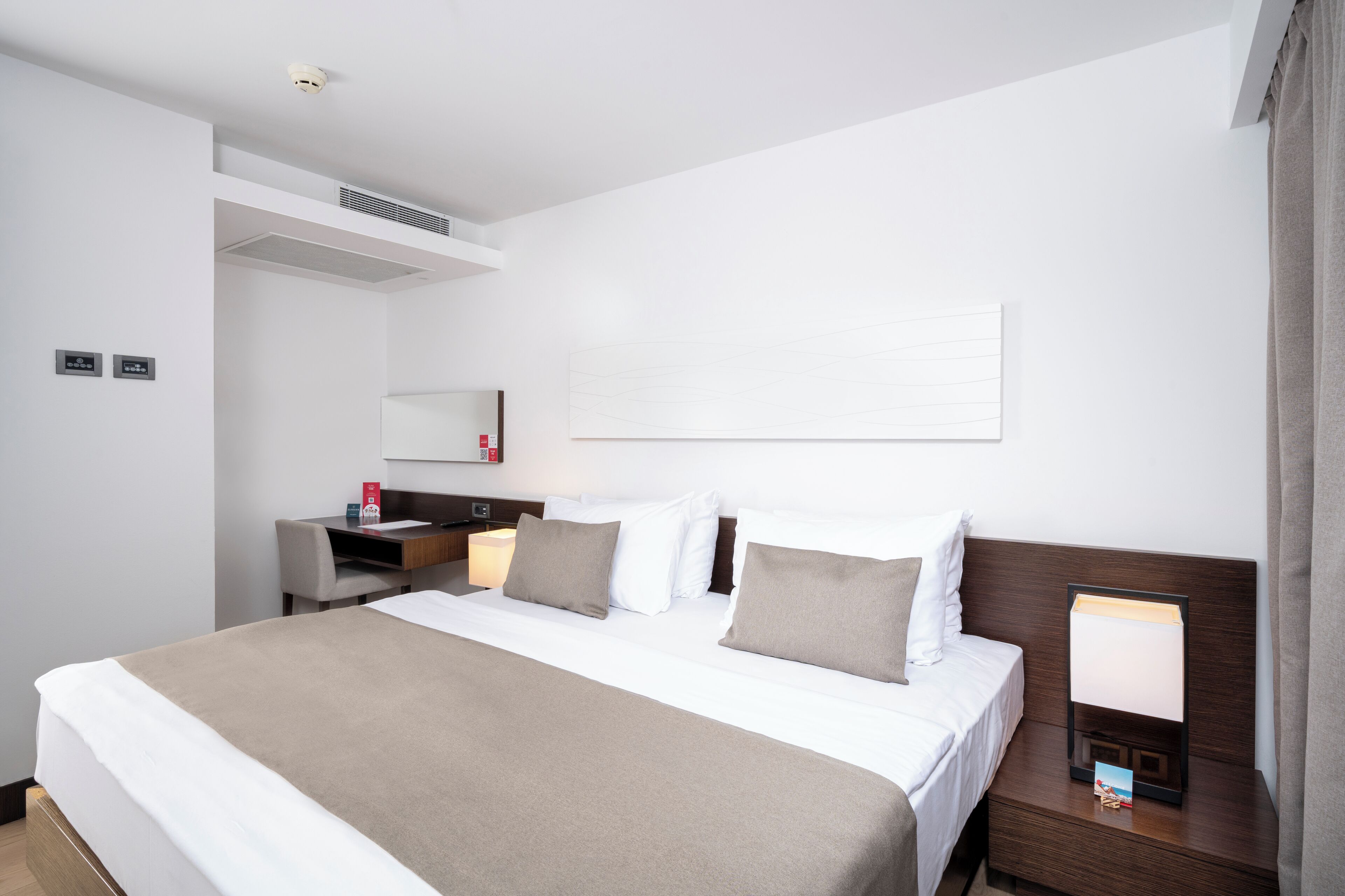 Habitación de hotel con una cama grande, un escritorio, una silla, una mesita de noche con lámpara y un estante en la pared.