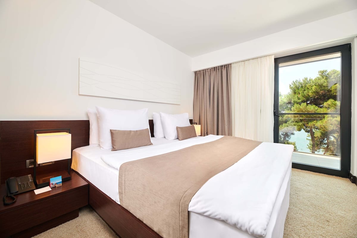 A hotel room with a bed, bedside table, lamp, and a view of trees through a large window.