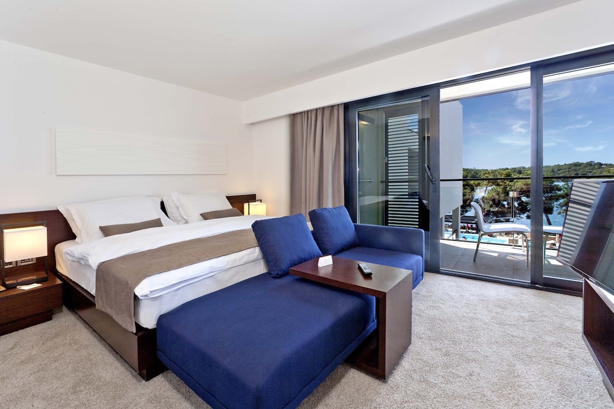 A modern hotel room with a bed, sofa, and a balcony with a pool view.