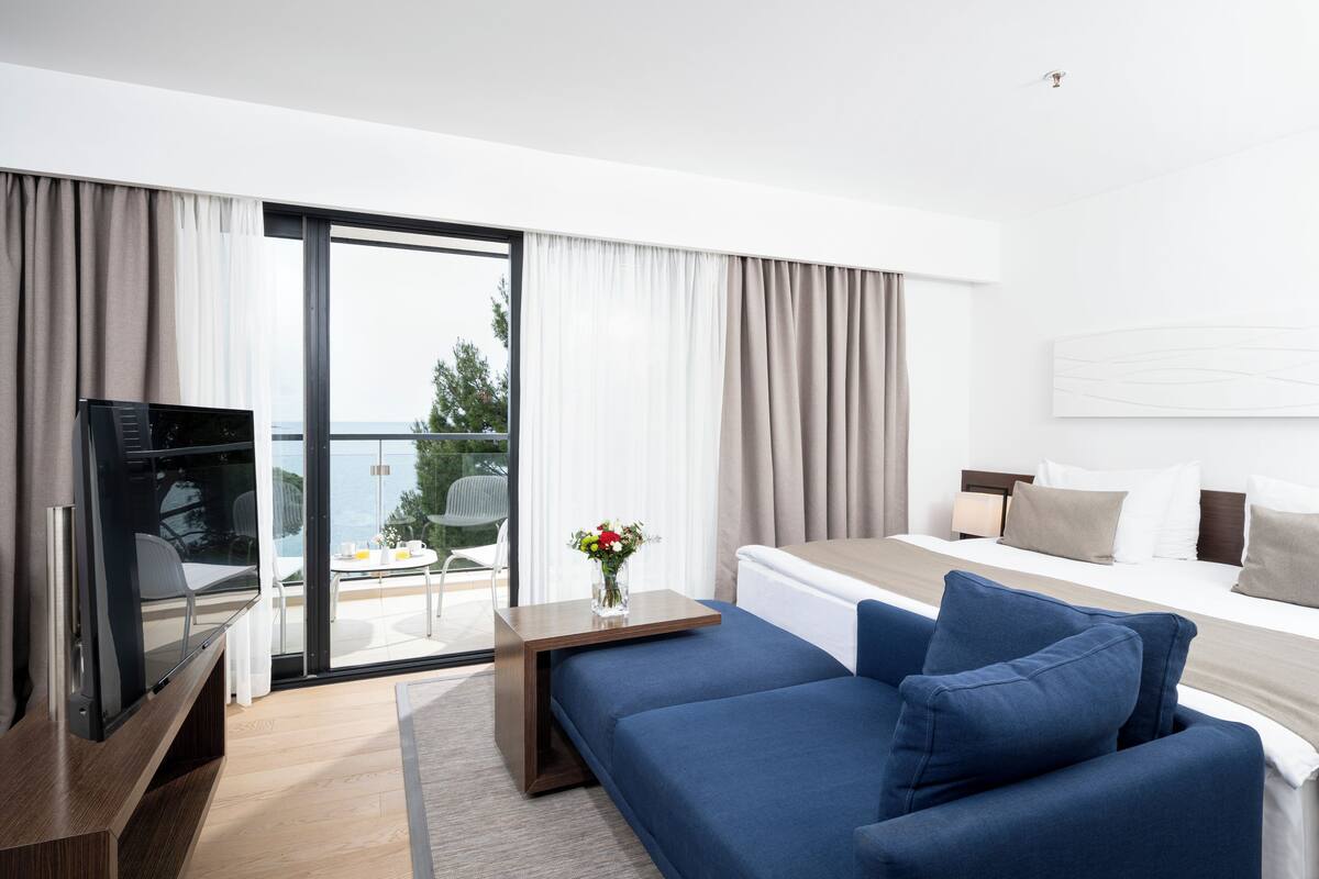 A modern hotel room with a bed, a blue sofa, a TV, and a balcony with a view.
