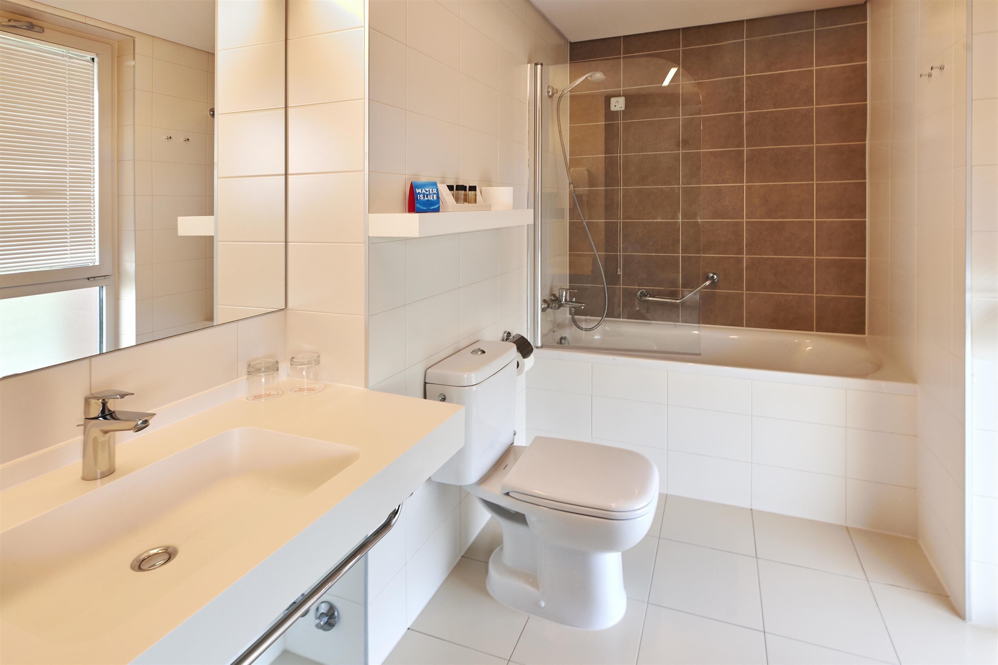 Un baño moderno con un lavamanos, inodoro y bañera blancos. Se observa un cabezal de ducha y una ventana con persianas.