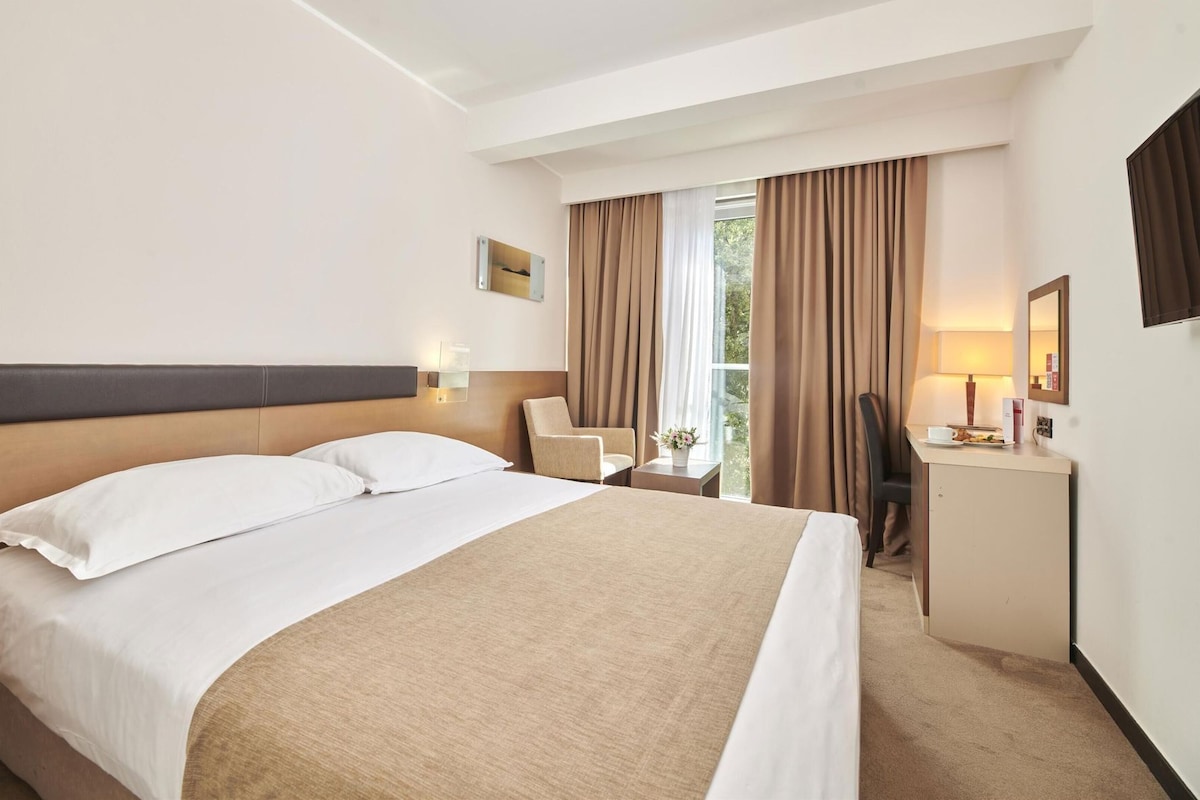 Una habitación de hotel con cama, escritorio, silla, lámpara y ventana con cortinas.