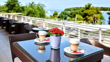 A table with two cups of coffee and a vase of red flowers on a balcony overlooking a scenic view.